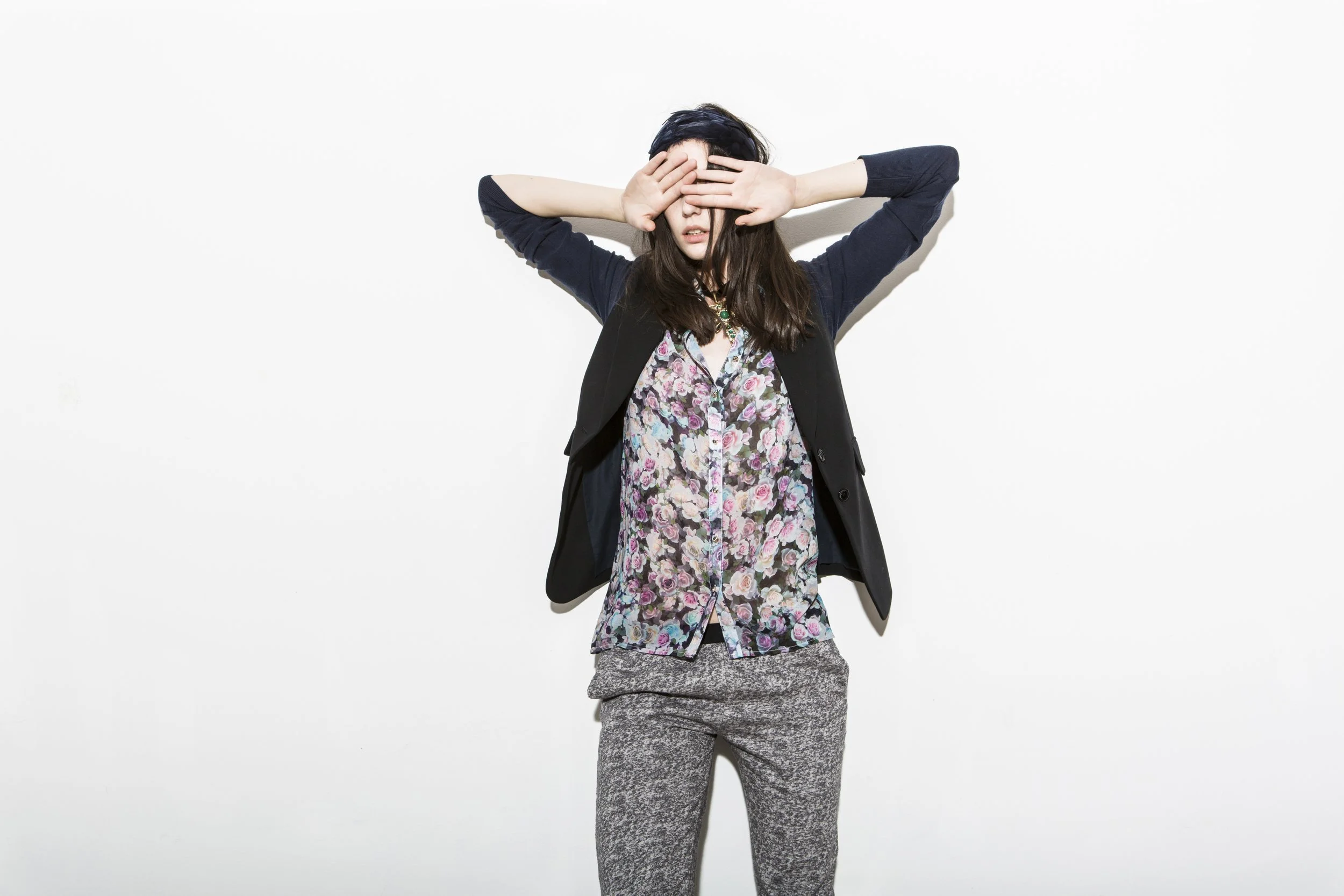 Une jeune femme pose devant un mur blanc, portant une veste noire, un chemisier floral, et un pantalon grisé de texture variée. Elle couvre ses yeux avec ses mains dans une posture expressive.