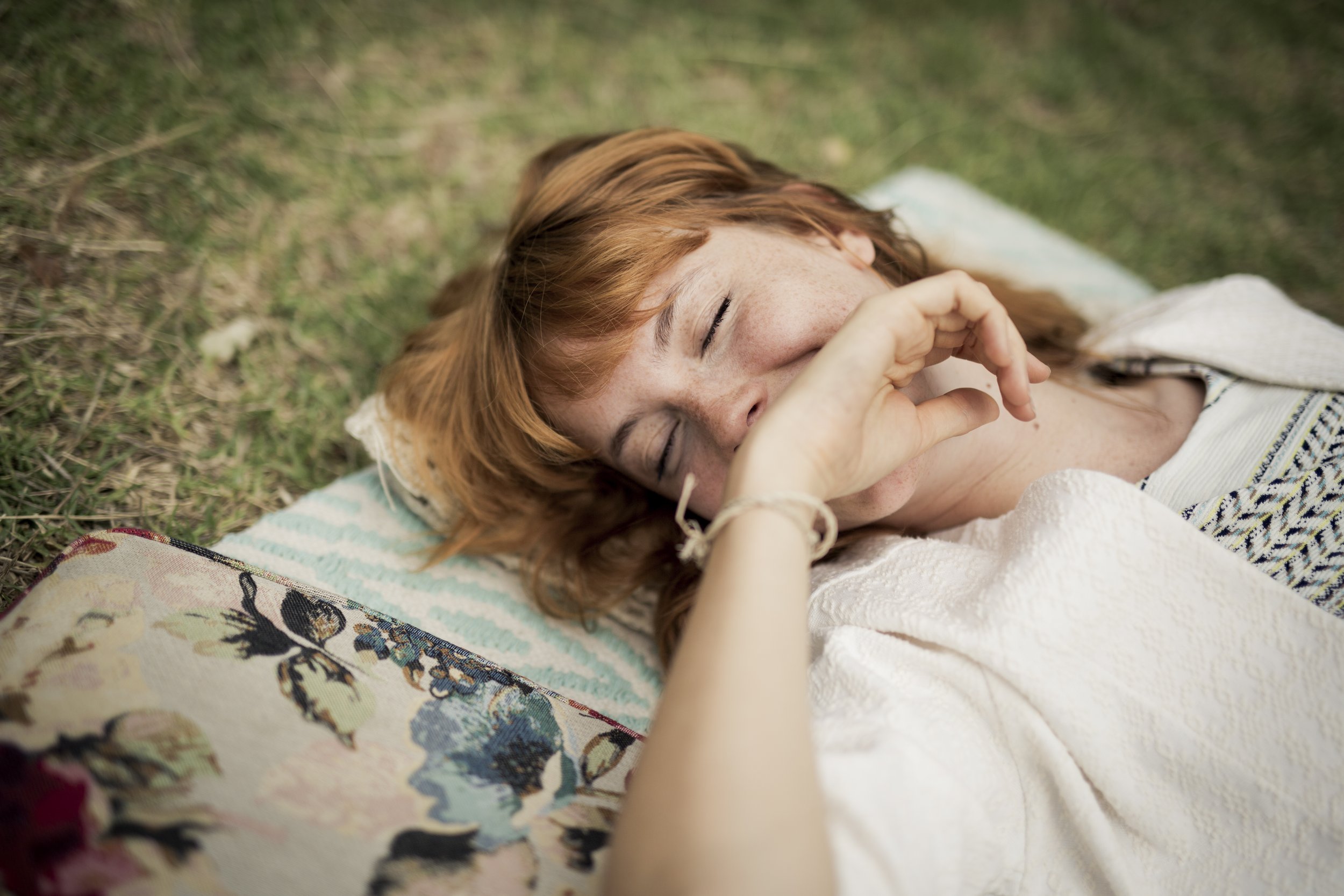 Une jeune femme aux cheveux roux repose sur l'herbe, éclatant de rire, avec un panier fleuri visible en premier plan.