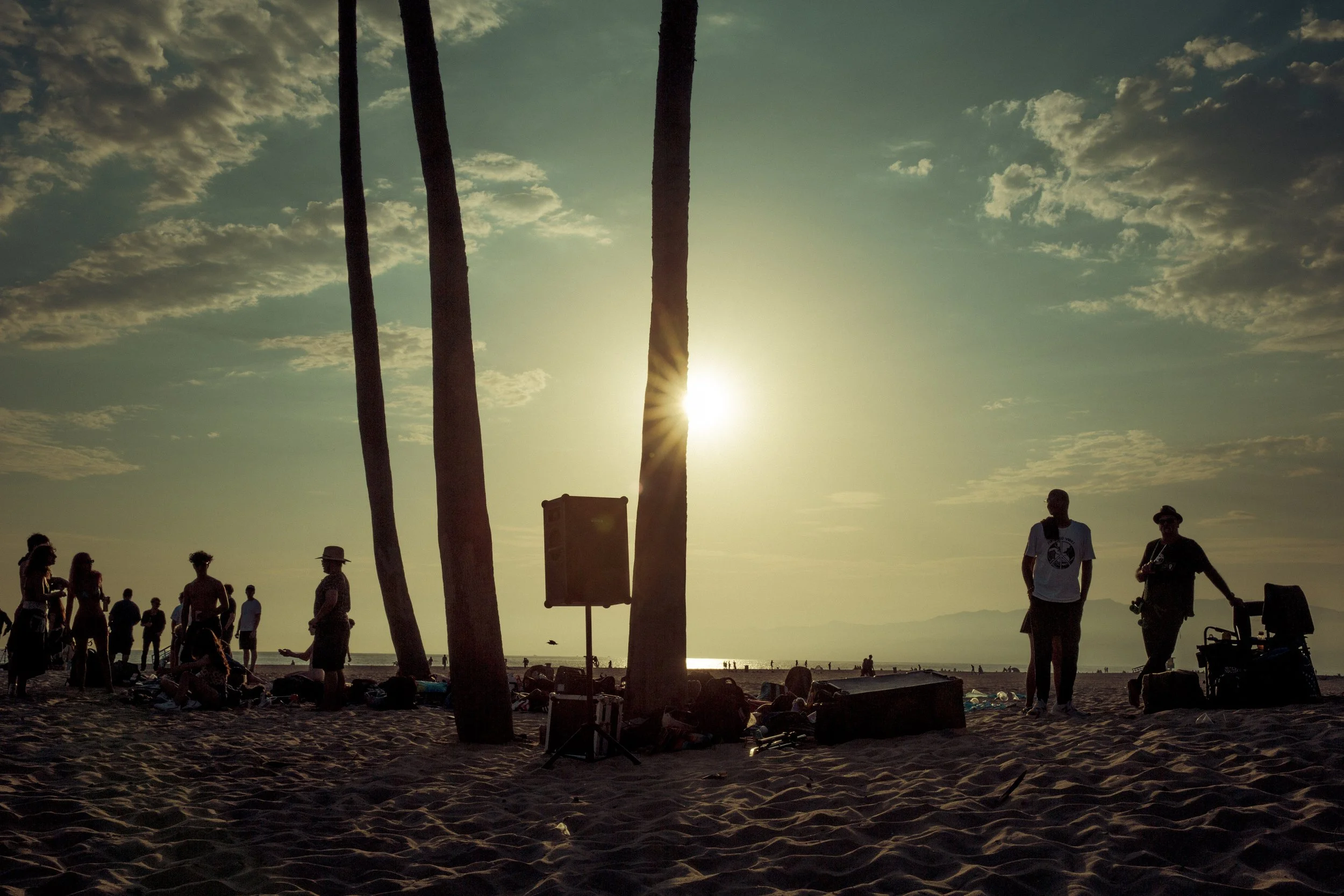 Scène de plage au coucher du soleil avec des silhouettes de personnes, des palmiers, et un ciel nuageux.