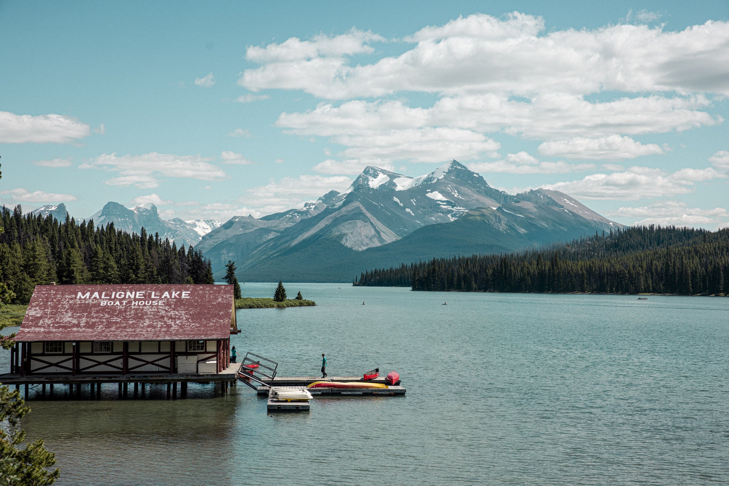 Lac Maligne avec montagnes enneigées en arrière-plan, forêt dense en premier plan, un bateau au quai et un bâtiment rouge avec une pancarte indiquant 'Maligne Lake Boat House'.