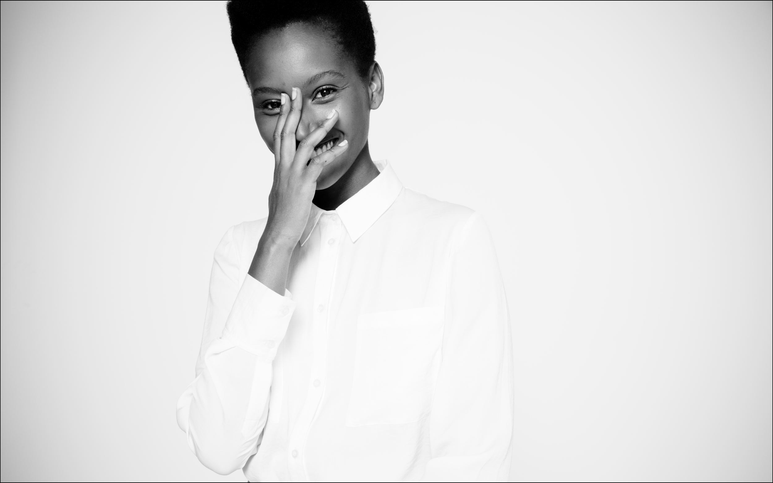 Photo en noir et blanc d'une femme souriante, portant une chemise blanche, avec la main gauche couvrant le visage partiellement, fond uni