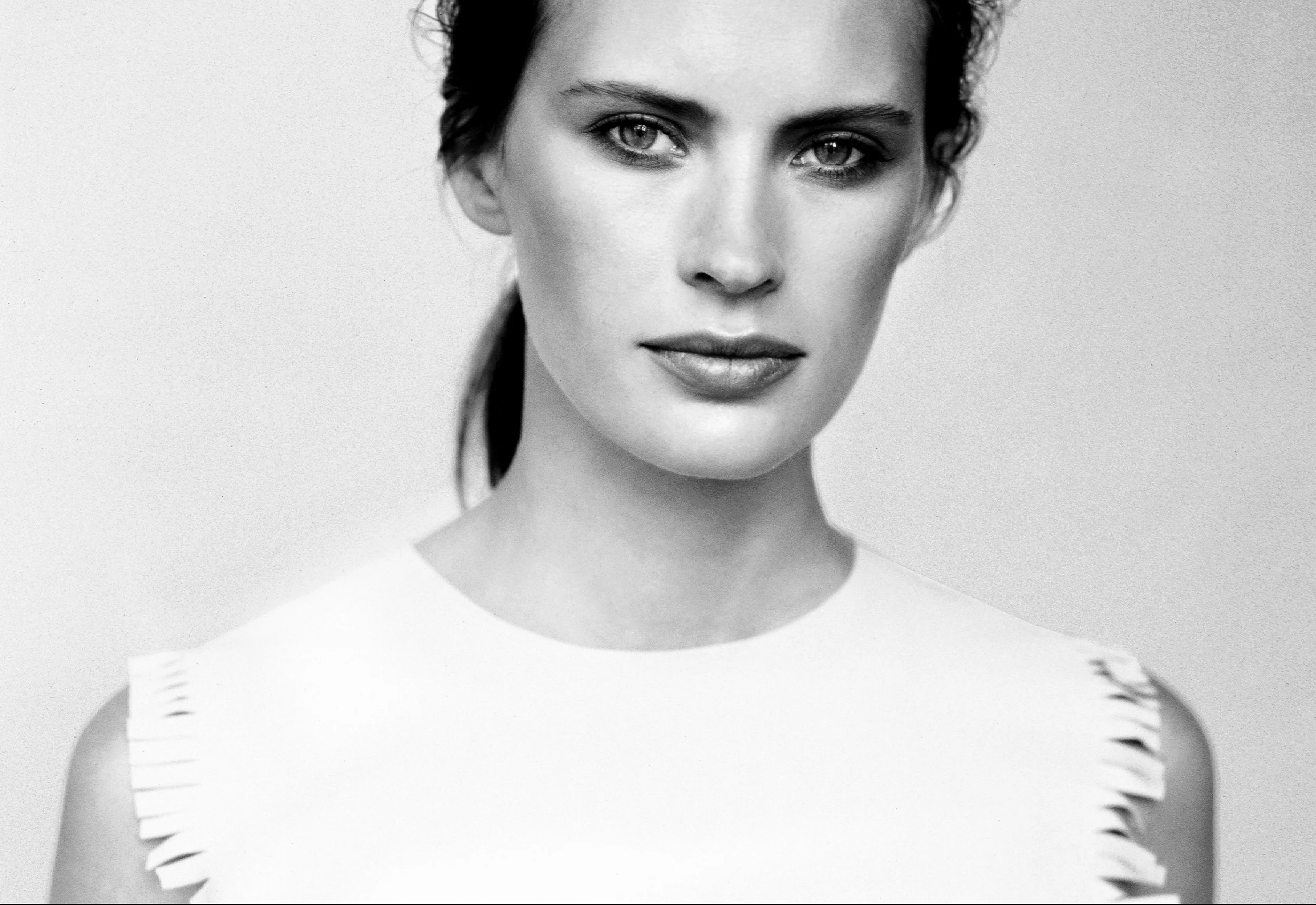 Un portrait en noir et blanc d'une femme avec des cheveux foncés, portant un haut moulant blanc avec des détails en découpe sur les épaules. La femme a une expression sérieuse et regarde directement l'objectif.