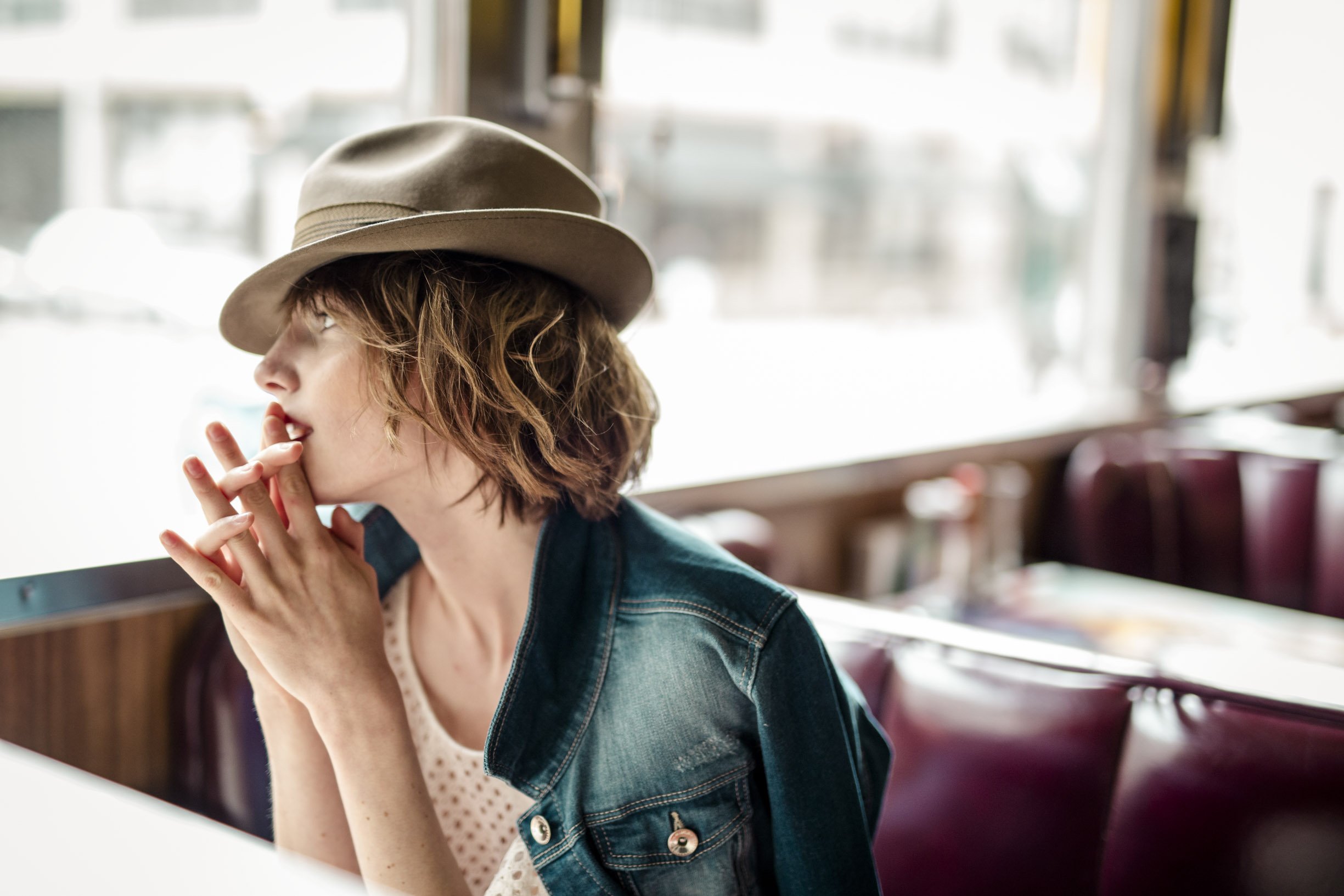 Une femme avec des cheveux bruns et courts, portant un chapeau beige et une veste en jean, assise dans un restaurant ou un café, en train de réfléchir ou de regarder dans une direction.