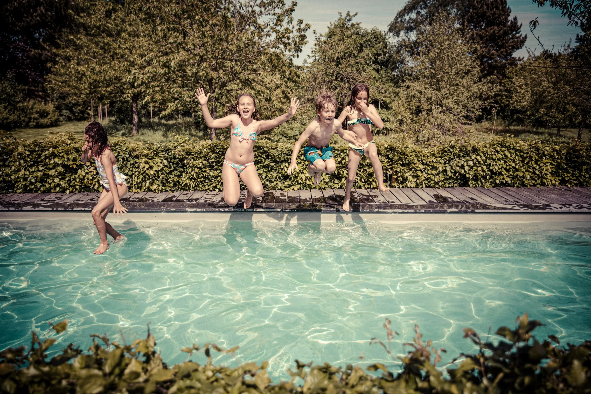 Quatre enfants sautent dans une piscine lors d'une journée ensoleillée, entourée de végétation verdoyante.