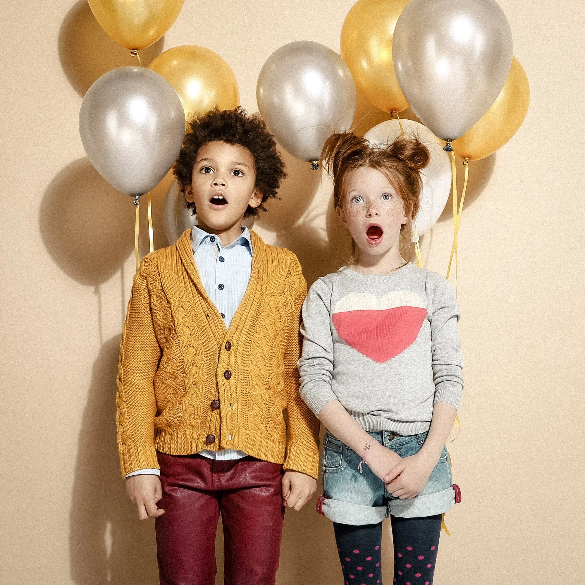 Deux enfants, un garçon aux cheveux frisés et une fille aux cheveux roux, debout devant un mur beige avec des ballons dorés, argentés et blancs derrière eux. La fille porte un sweat gris avec un cœur rose, et le garçon porte un cardigan épais orange 