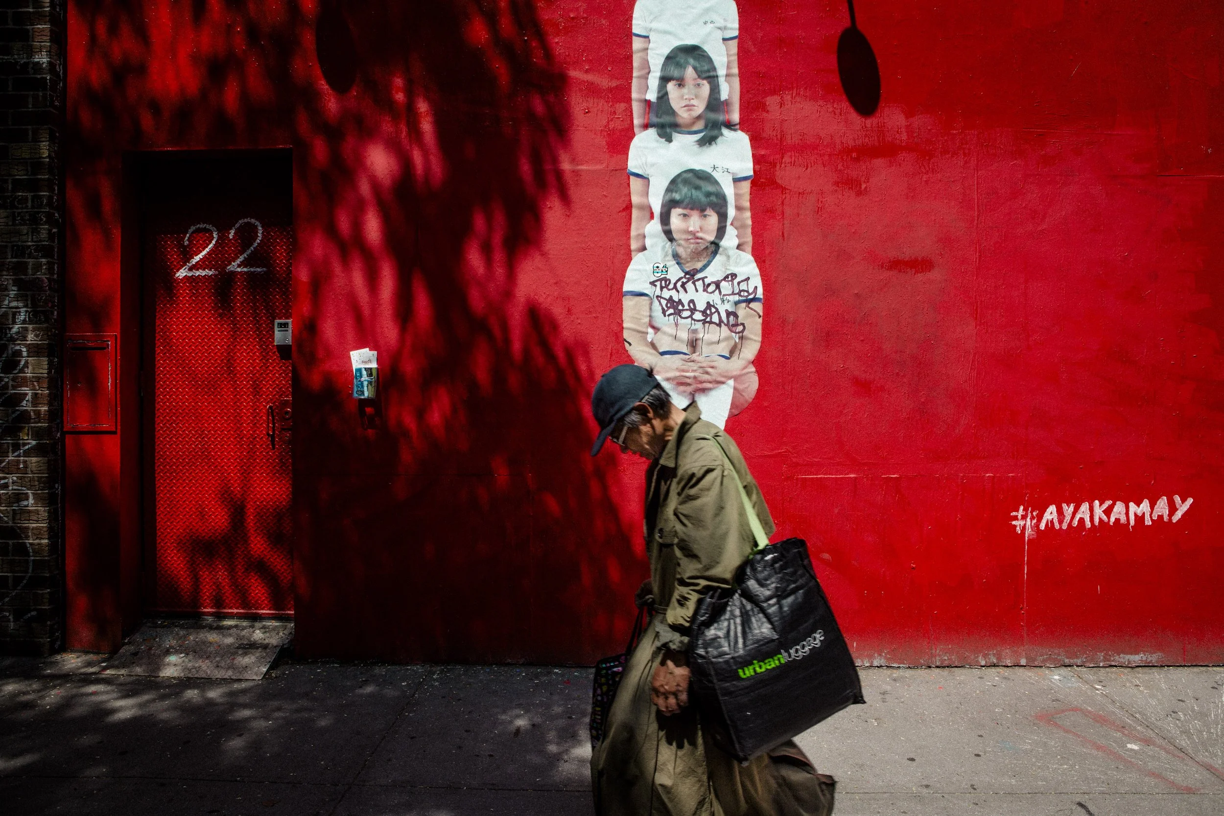 Une affiche murale rouge montre deux femmes avec des coiffures bob, une plus vieille et une plus jeune, en vêtements de style scolaire. Un homme portant un chapeau et un manteau beige marche devant, portant un sac noir avec la inscription 'urban' en 