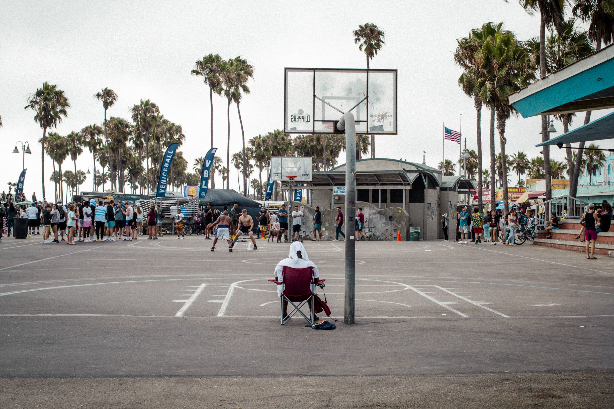 Une scène de plage en Californie avec de nombreux personnes jouant au basketball près de terrains de sport, des palmiers, un drapeau américain, et des stands et bâtiments colorés dans un environnement de bord de mer.