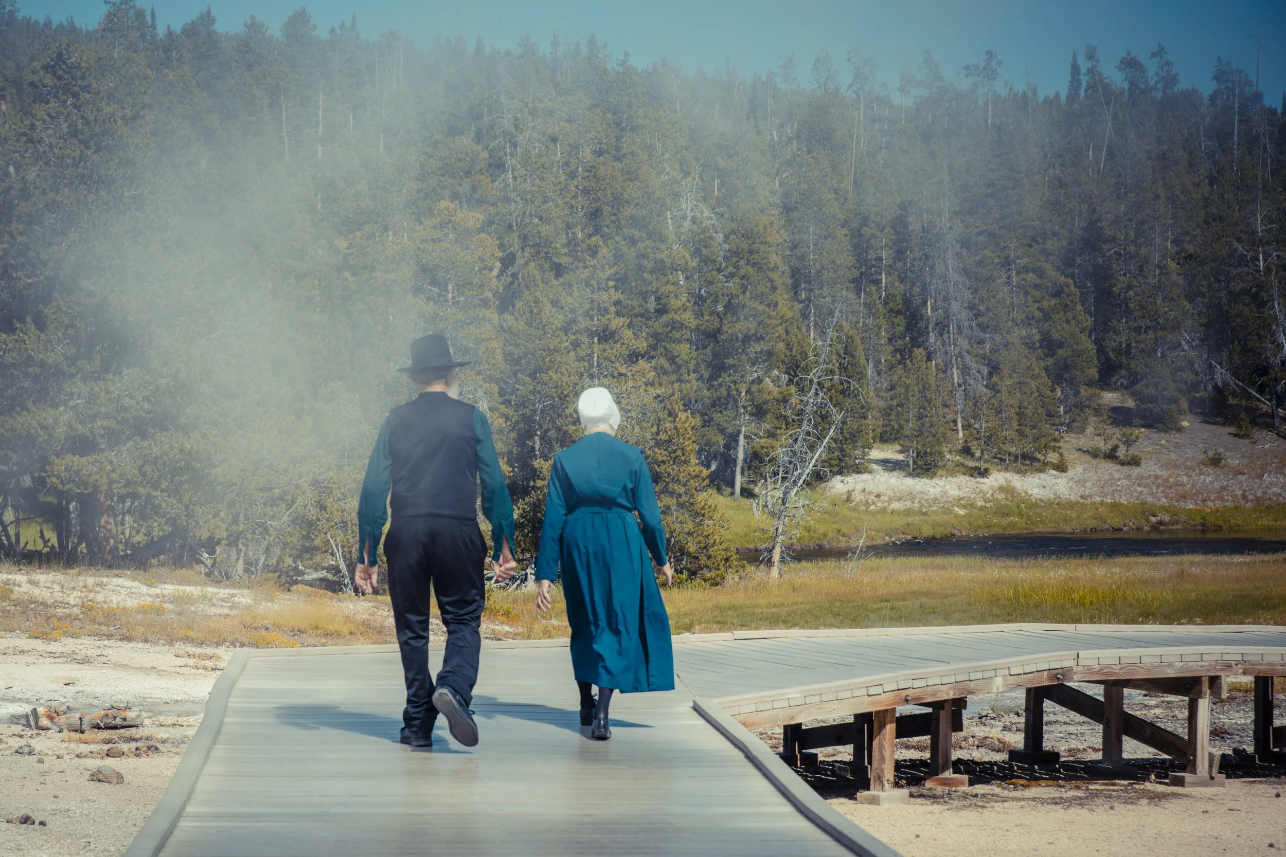 Deux personnes âgées, un homme et une femme, marchent main dans la main sur un pont en bois dans un environnement naturel avec beaucoup d'arbres et un lac à proximité.