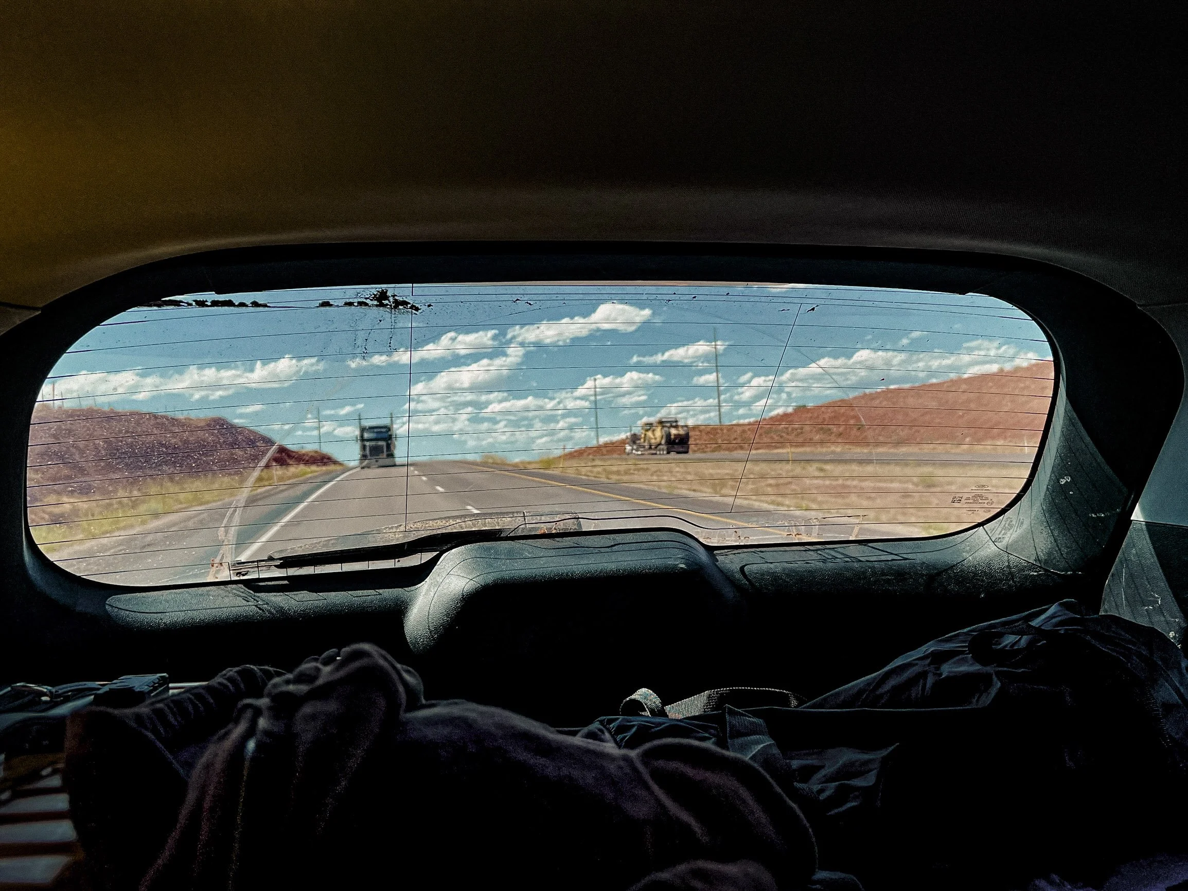 Vue depuis l'intérieur d'une voiture vers l'arrière, montrant une route en montée, un ciel bleu avec des nuages, et des véhicules en marche, avec des montagnes ou des collines en arrière-plan.