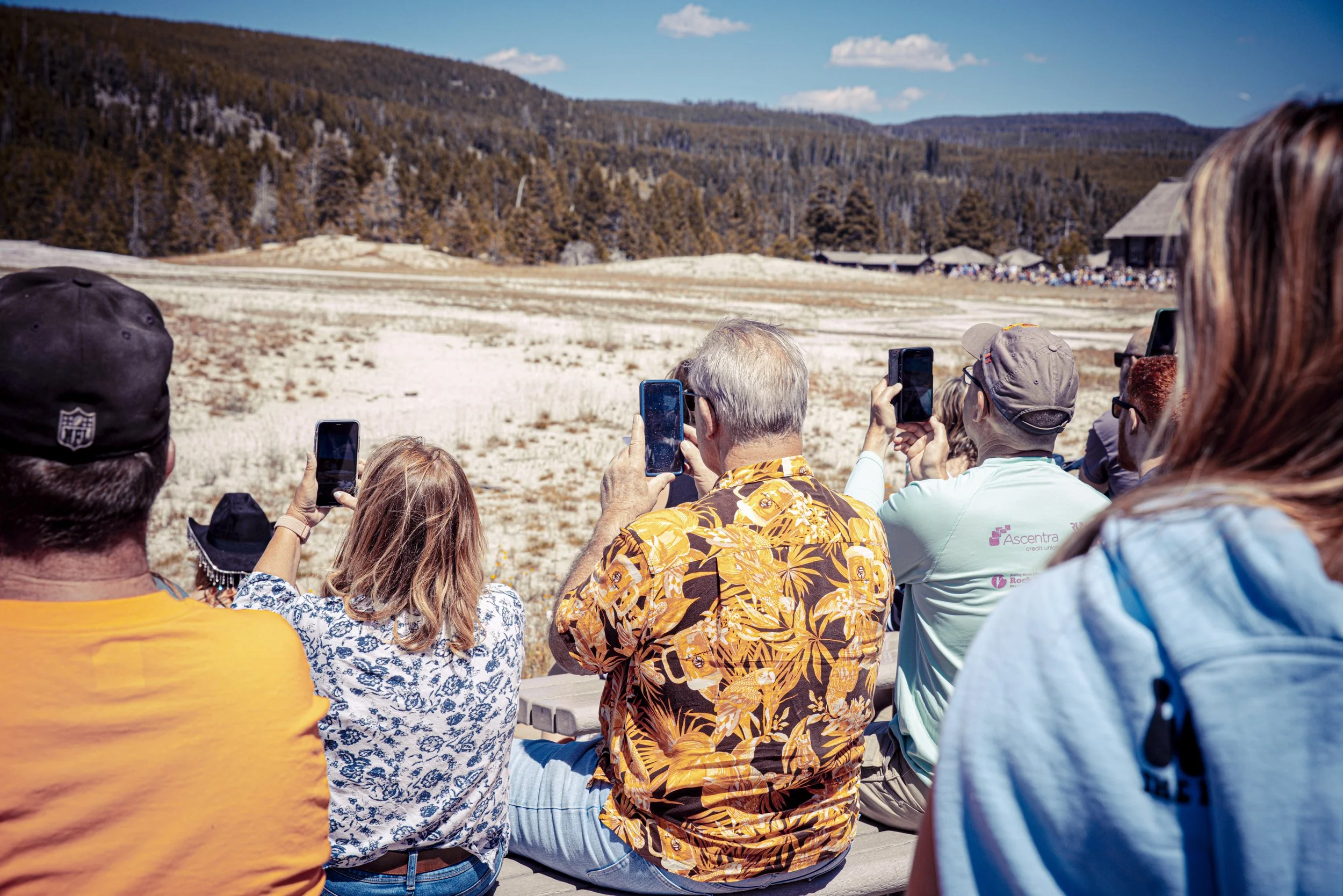 Groupe de personnes assises en plein air, regardant un paysage naturel avec des montagnes et des forêts, en prenant des photos avec leurs smartphones.