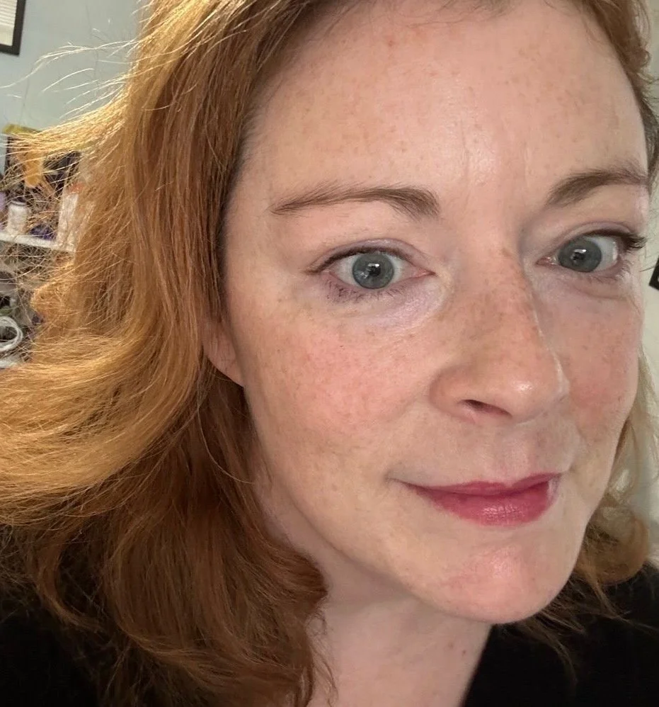 Close-up of a woman with red hair and blue eyes, smiling slightly, with freckles on her face.