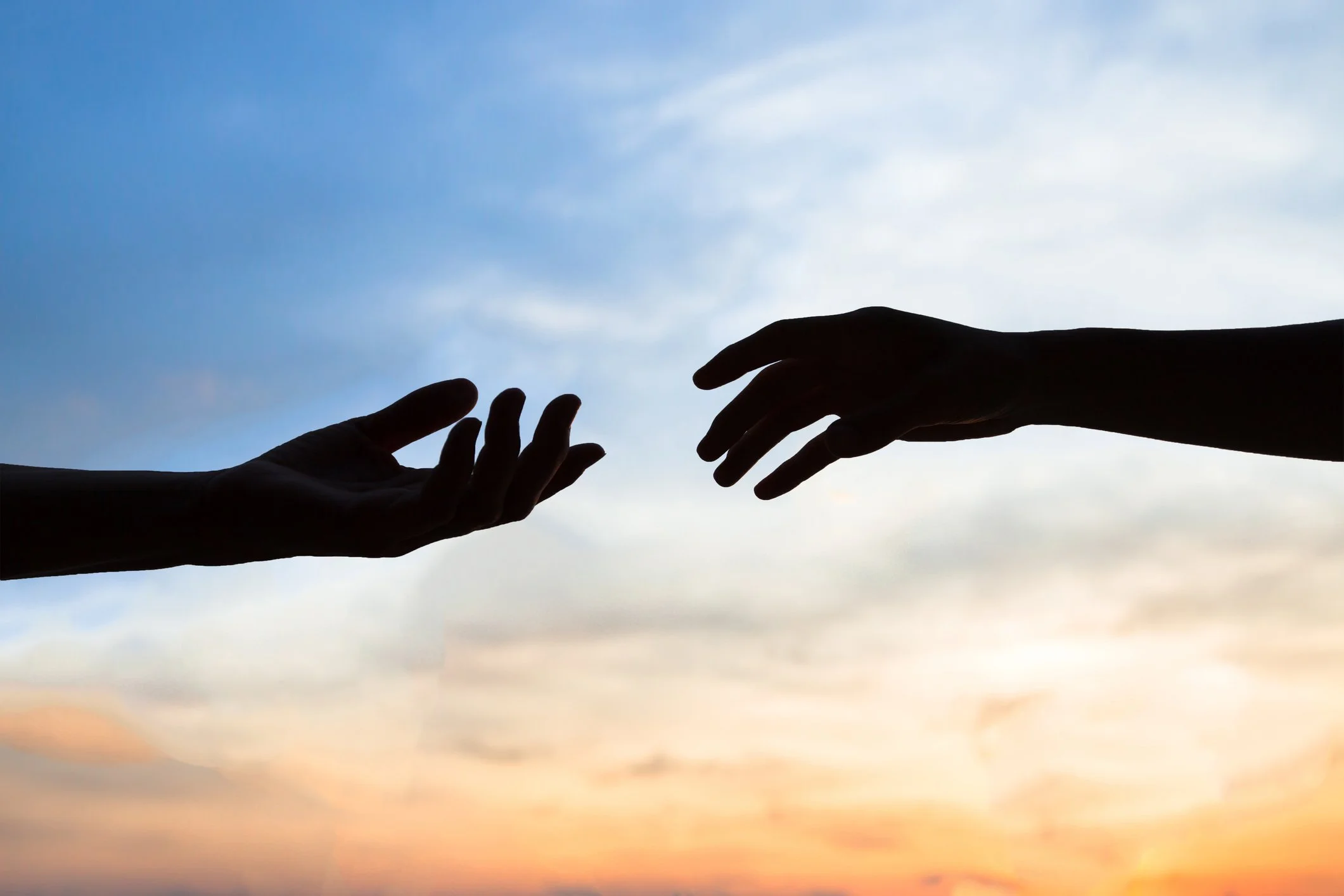 Two hands reaching towards each other against a sky with clouds and a sunset.