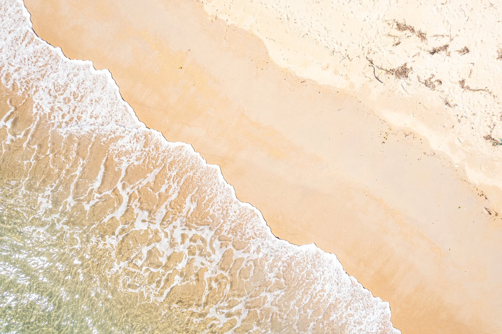 Aerial view of a sandy beach with waves washing onto the shore and seaweed scattered along the sand.