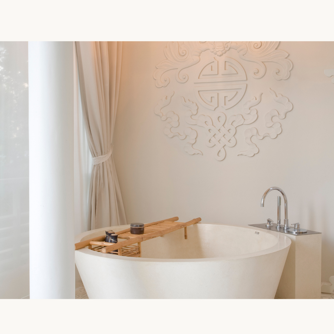 A luxurious bathroom featuring a modern oval bathtub with a wooden bath tray, beige curtains, and a decorative wall sculpture with intricate designs.
