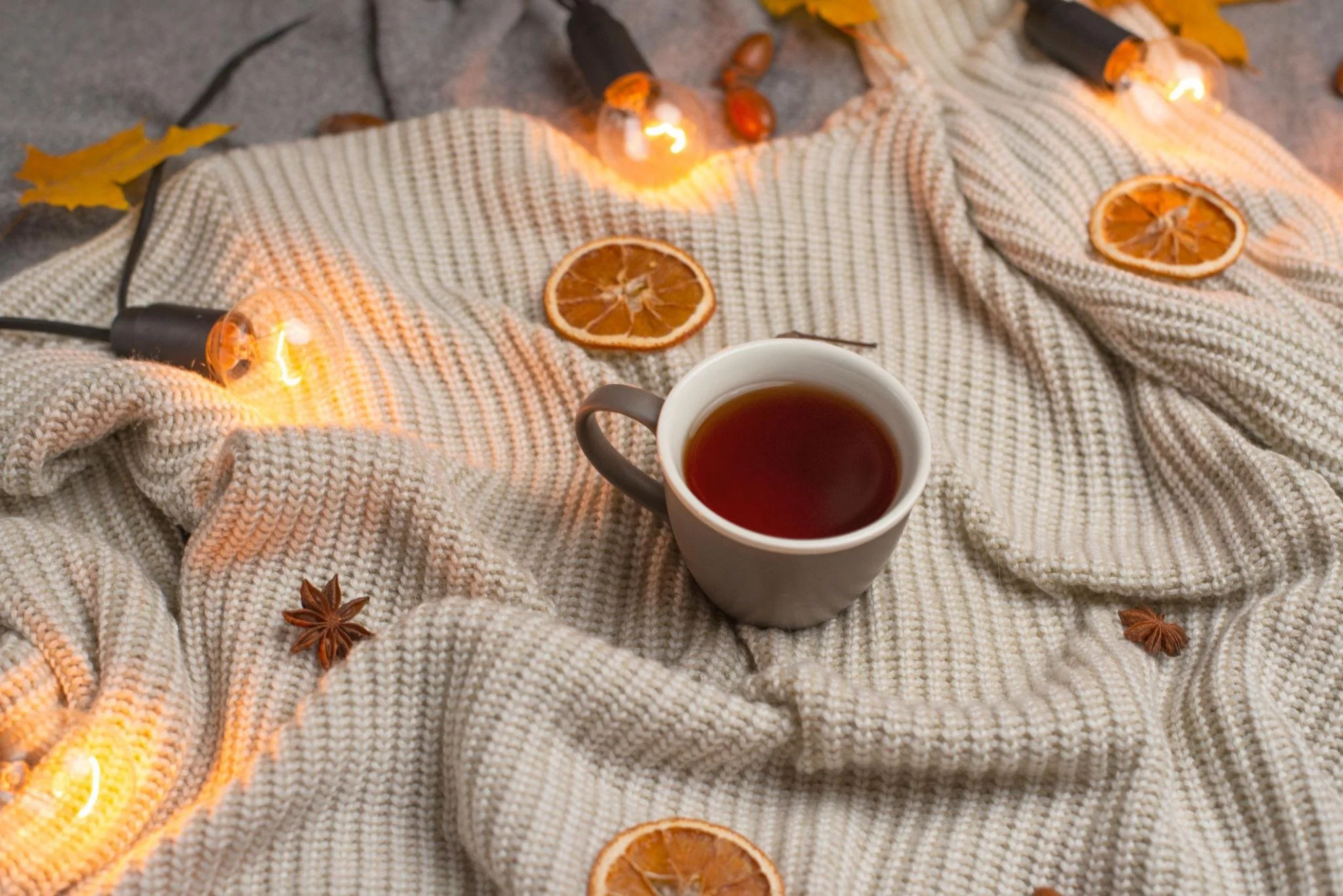 Une tasse de thé sur une couverture en laine avec des tranches d'orange séchées, des branches d'anis étoilé, une guirlande lumineuse et des feuilles d'automne.