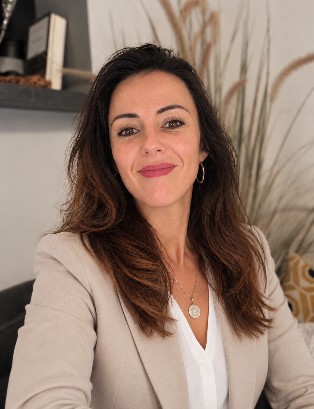 Portrait d'une femme souriante avec des cheveux bruns ondulés, portant un blazer beige, une veste blanche, un collier et des boucles d'oreilles, posant dans un intérieur décoré de plantes et d'objets décoratifs.