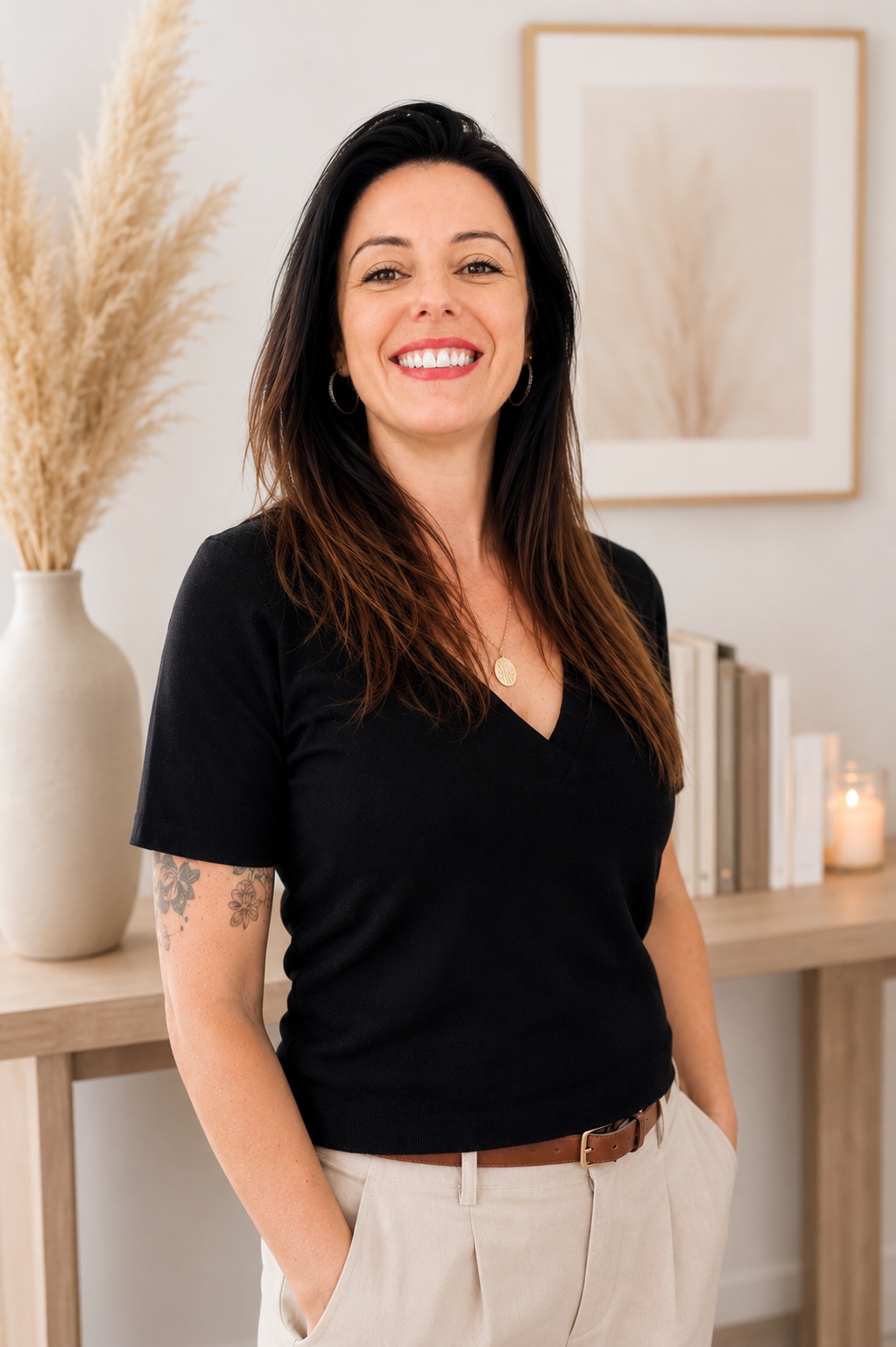 Une femme souriante avec des cheveux bruns longs, portant un t-shirt noir et un pantalon beige, se tenant dans une pièce décorée avec un vase de pampas et des livres sur une table.