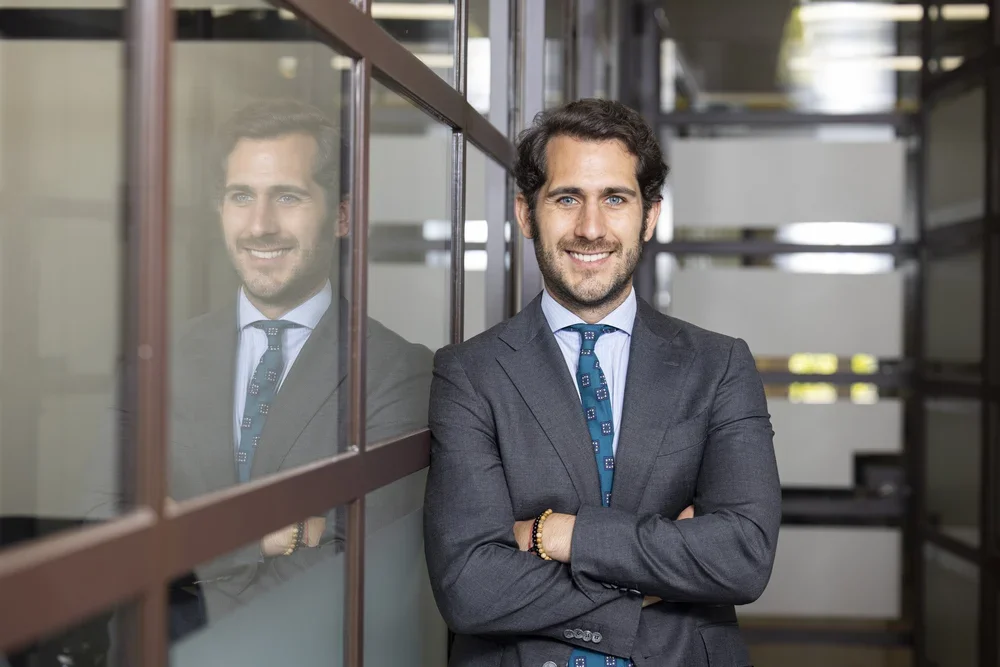 Hombre de negocios sonriendo, vestido con traje gris y corbata azul, de pie con los brazos cruzados junto a una pared de vidrio reflectante en un ambiente de oficina moderna.