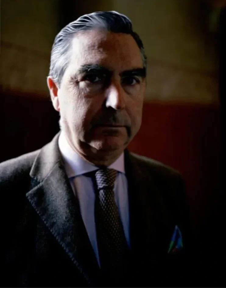 Retrato de un hombre con cabello gris y bien peinado, usando un saco oscuro, camisa blanca y corbata, en un ambiente de interior con iluminación suave.