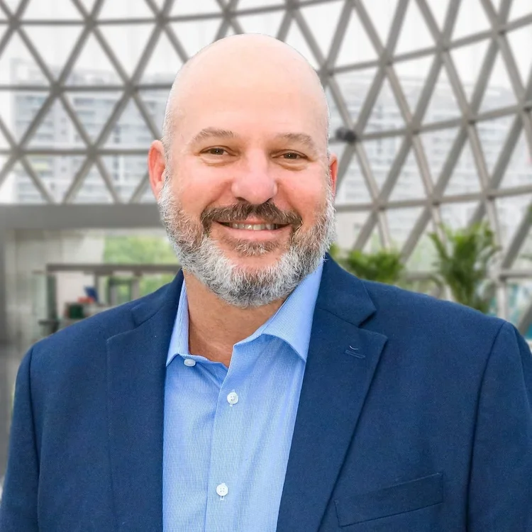 Hombre calvó con barba y camiseta azul, usando chaqueta azul, sonriendo en un entorno moderno con estructura de vidrio y plantas.