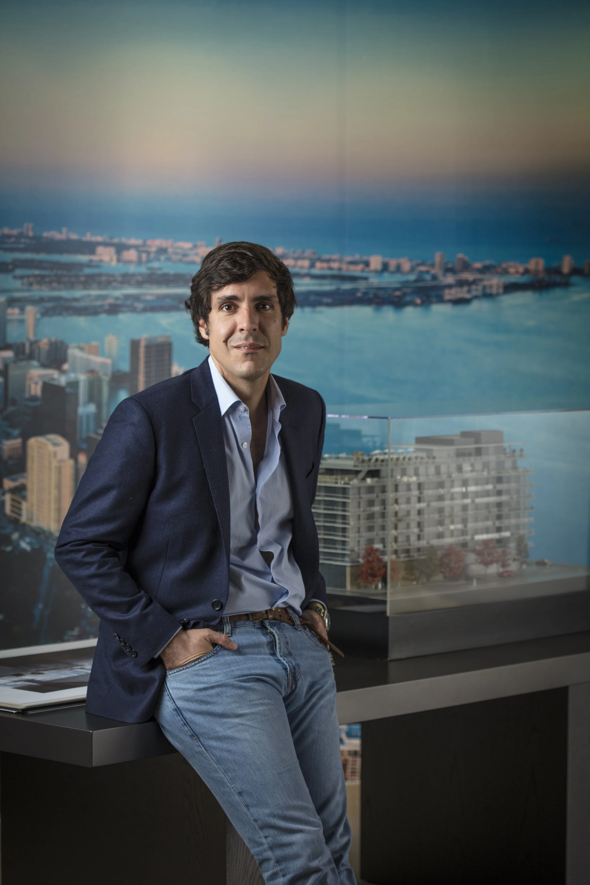 Hombre con cabello oscuro y sudadera azul en una oficina con vista panorámica de la ciudad y el mar.