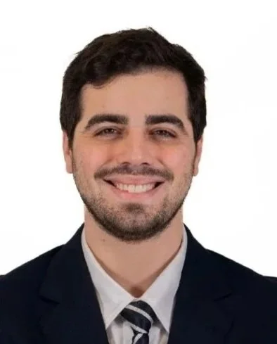 Joven hombre con barba, sonriendo, usando traje y corbata, con fondo blanco.