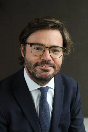 Retrato de un hombre con gafas, barba y cabello oscuro, vestido con saco y corbata, sonriendo frente a un fondo gris.