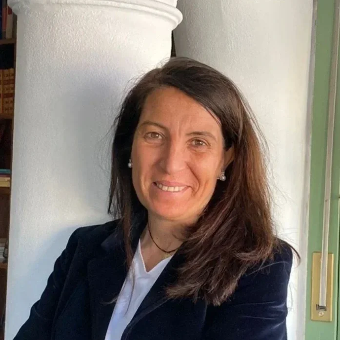 Retrato de una mujer con cabello castaño oscuro, sonriendo, vestida con chaqueta negra y blusa blanca, en un entorno interior
