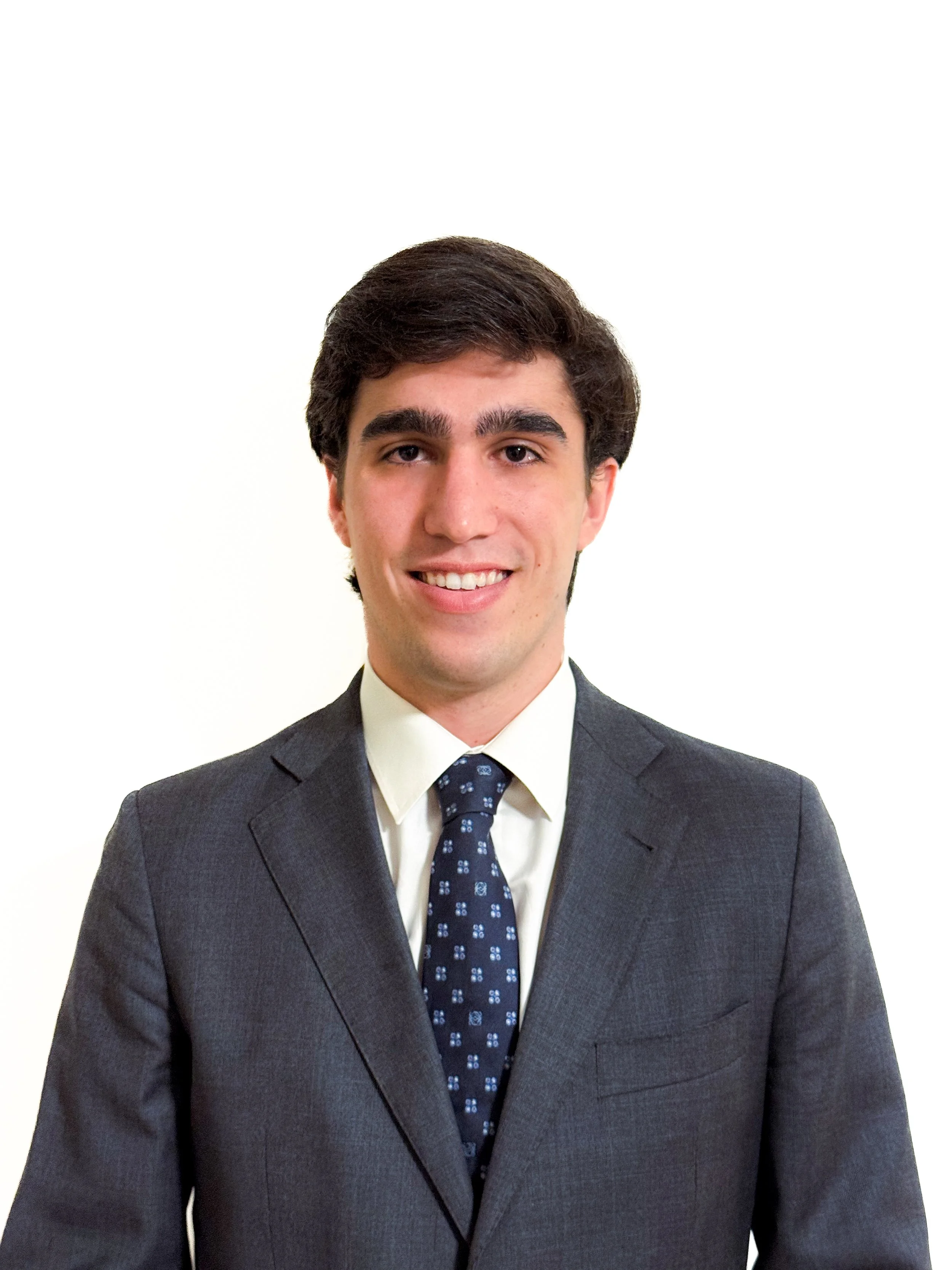 Joven hombre con cabello castaño, vistiendo saco gris, camisa blanca y corbata azul con patrones blancos, sonriendo.