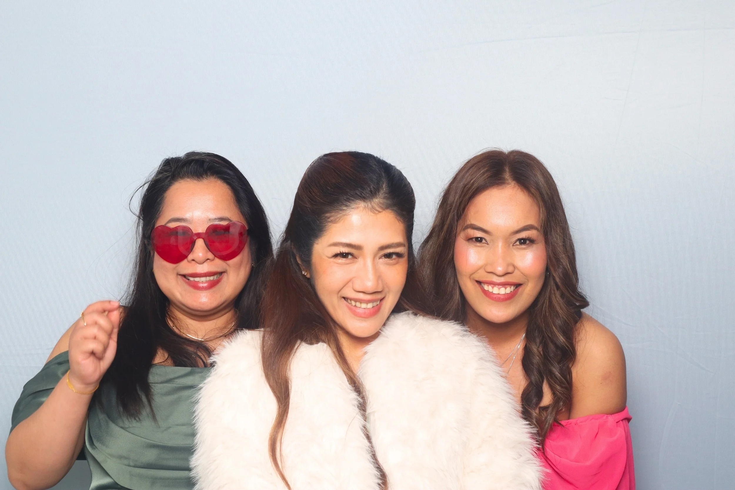 Winter Wedding guests posing together in front of the premium background; woman on the left wears red heart-shaped sunnies, woman in the middle wears a white fur coat, and woman on the right wears a pink off shoulder top. Flawless raw pictures