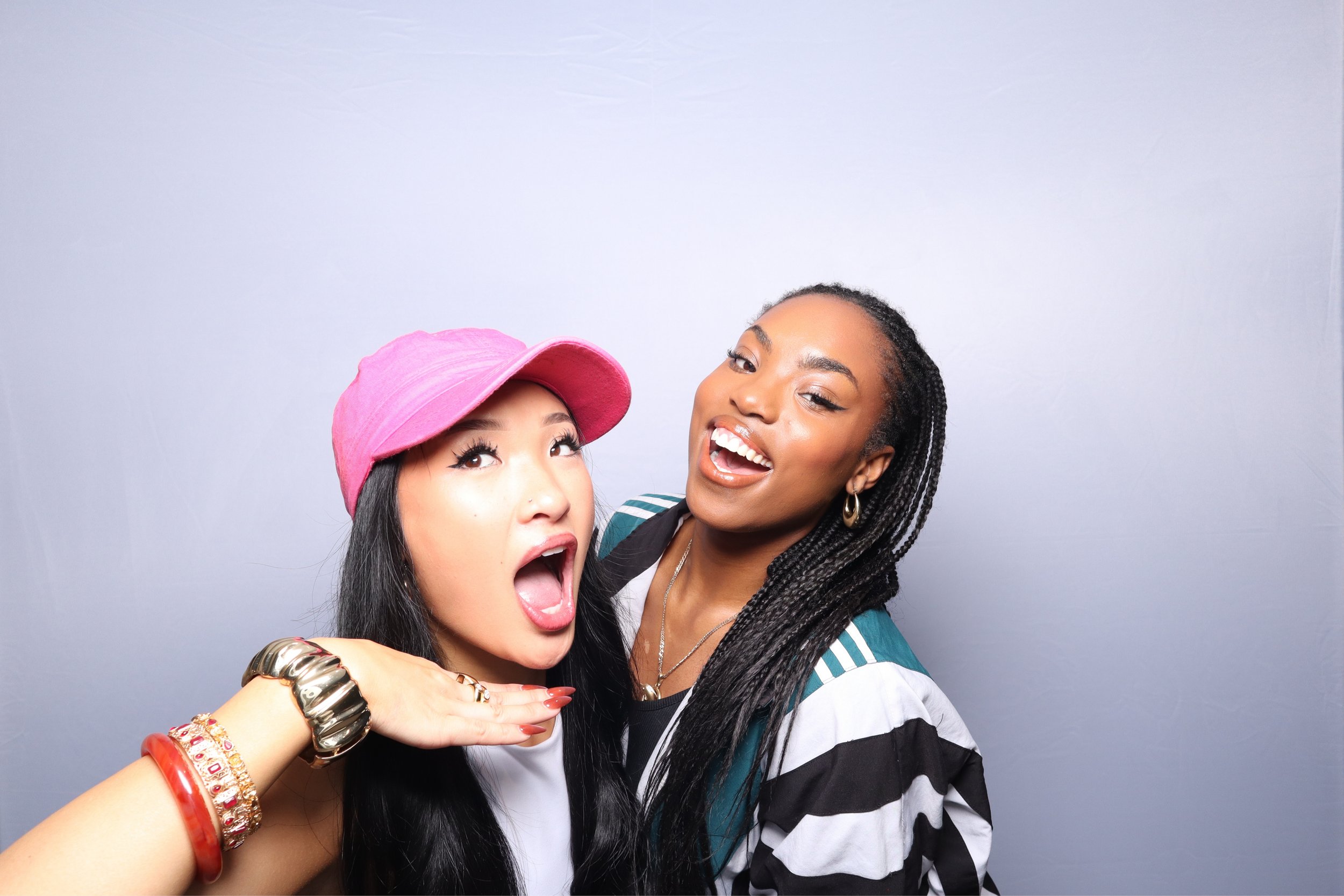 Two pretty stylish women smiling and posing against a grey background. One woman has braided hair, earrings, and is wearing a striped jacket. The other woman has long dark hair, pink hat, and multiple colorful bracelets. KRANIRAIVU event fashion show