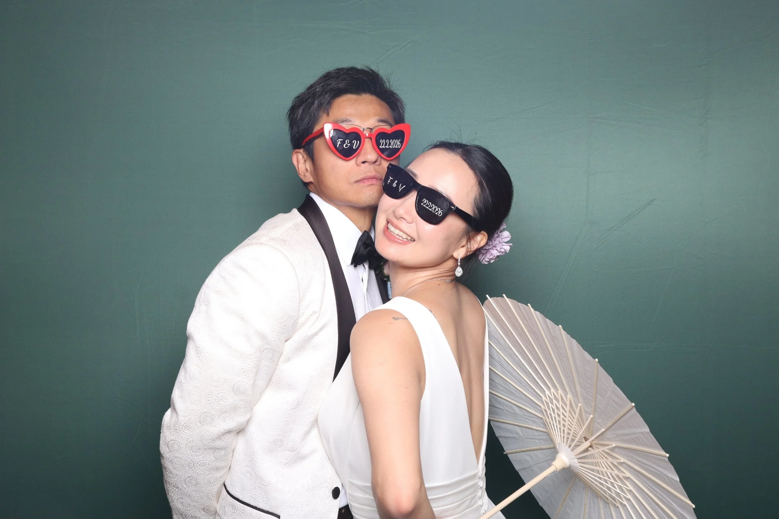 Elegant Bride & groom wearing customised sunglasses from the booth. The man is wearing a white suit tuxedo and the bride is radiant, smiling, holding a white parasol. They are standing against a green backdrop floral theme. 