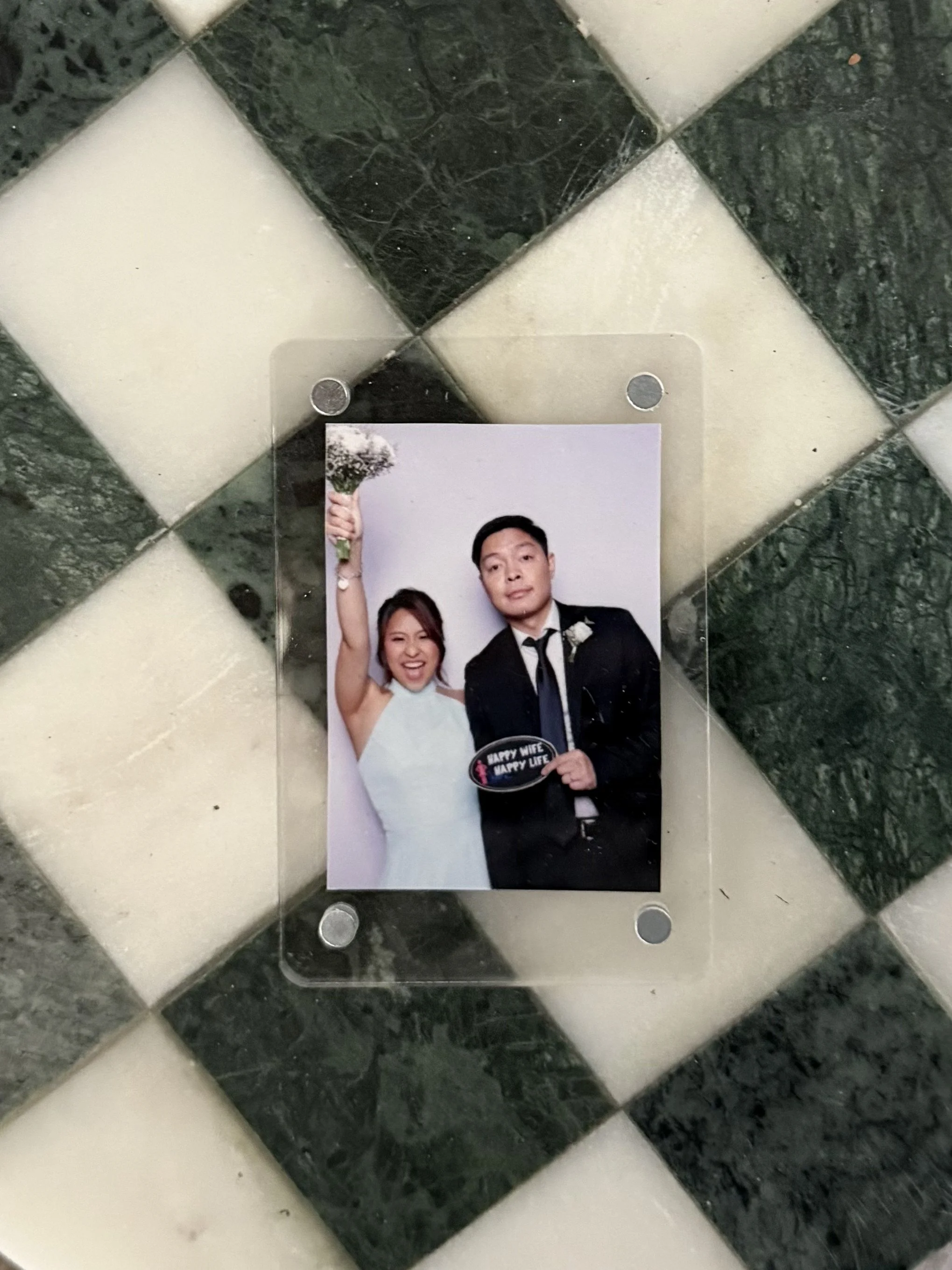 Photo of a wedding photo in a clear frame on a black and white checkered marble floor. The photo shows a couple in formal attire, with the woman smiling and the man holding a sign that says "Happy Wife, Happy Life". The woman is wearing a white dress and the man is wearing a black suit.
