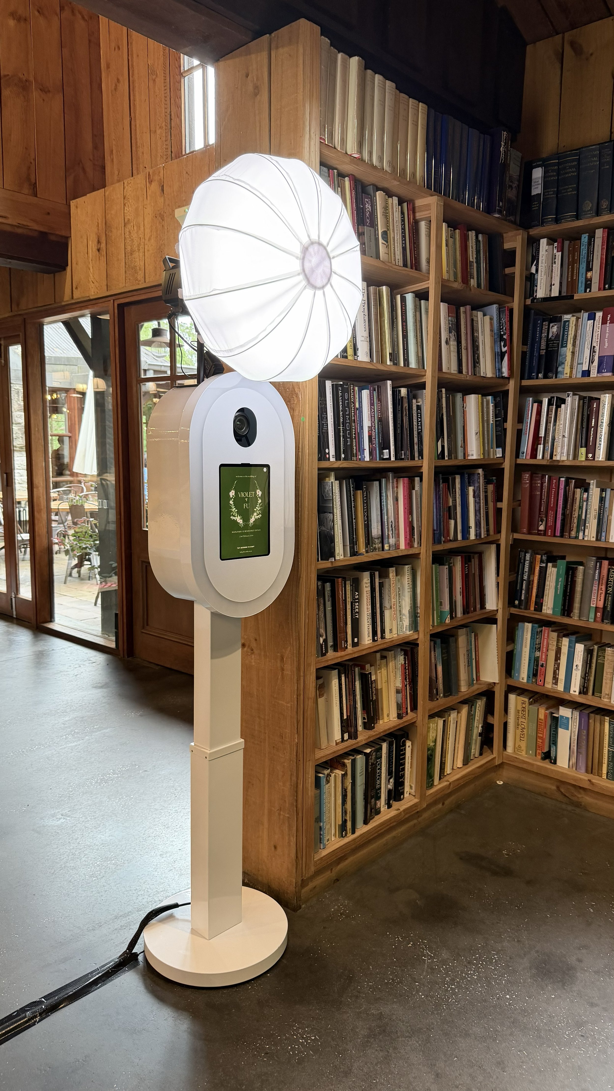 Photo of a digital photo booth with a ring light and a screen displaying 'Violet Fu' inside a bookstore or library with wooden bookshelves filled with books.