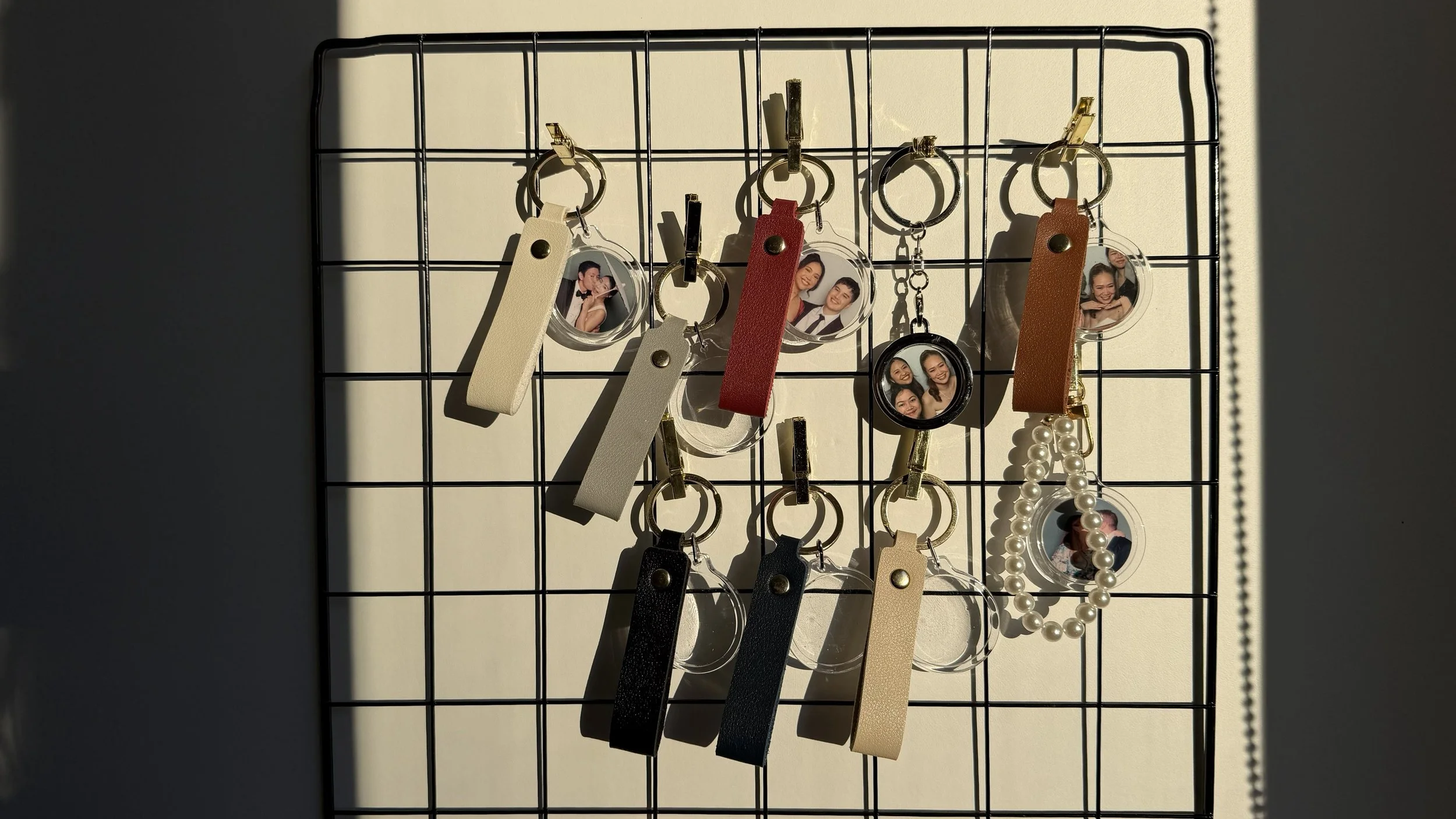 A grid display board with various keychains, photo charms, and accessories, some attached with black clips, illuminated by sunlight casting shadows.