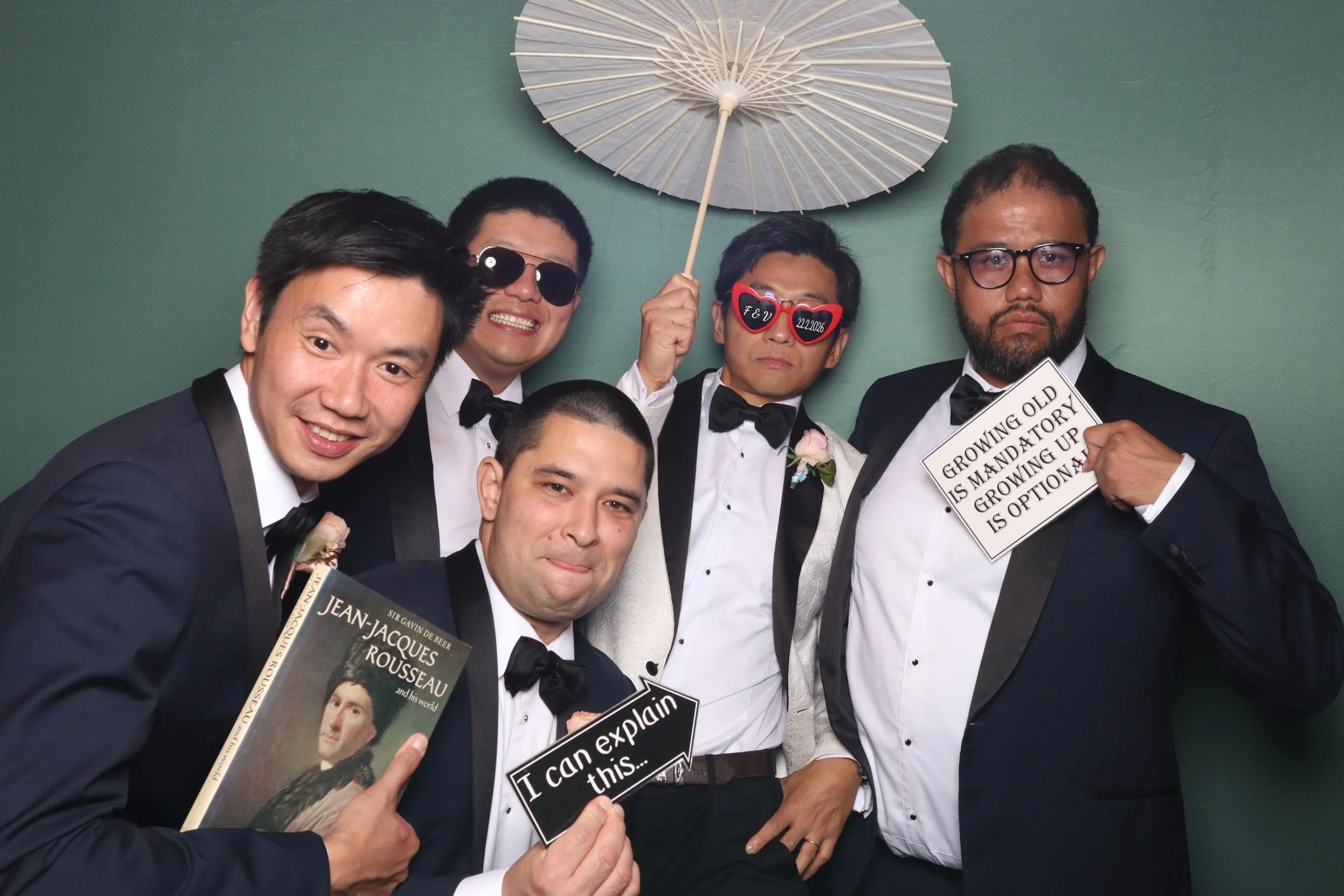 Group of groomsmen in tuxedos posing for three stills modern clean white photobooth holding book props, a parasol, and customised sunnies / glasses and props in. Garden floral theme at bookbarn intimate wedding