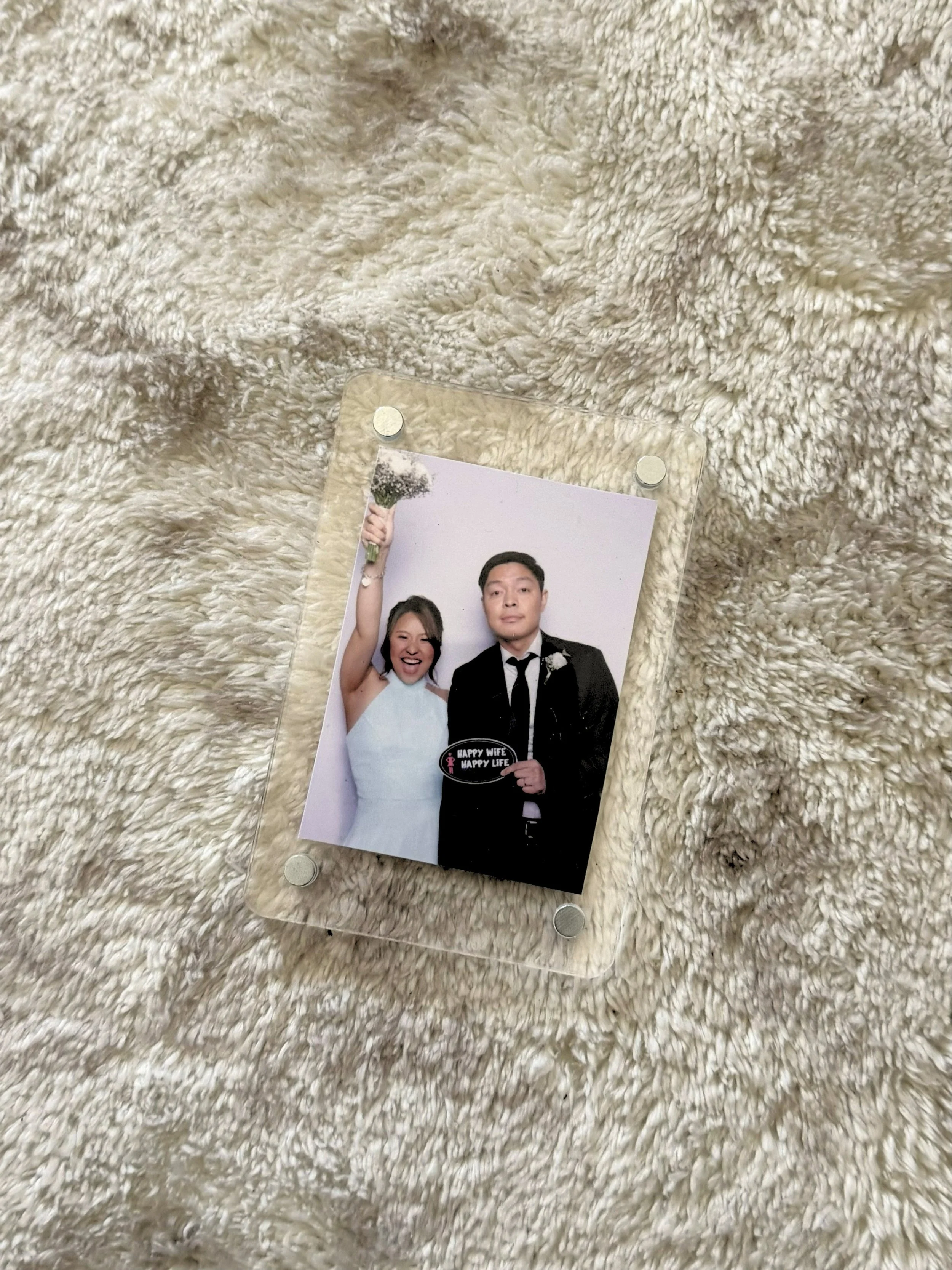 A wedding photo of a newlywed couple dressed in wedding attire, with the bride smiling and holding a bouquet, and the groom standing beside her. They are inside a photo frame on a fluffy, cream-colored textured surface.