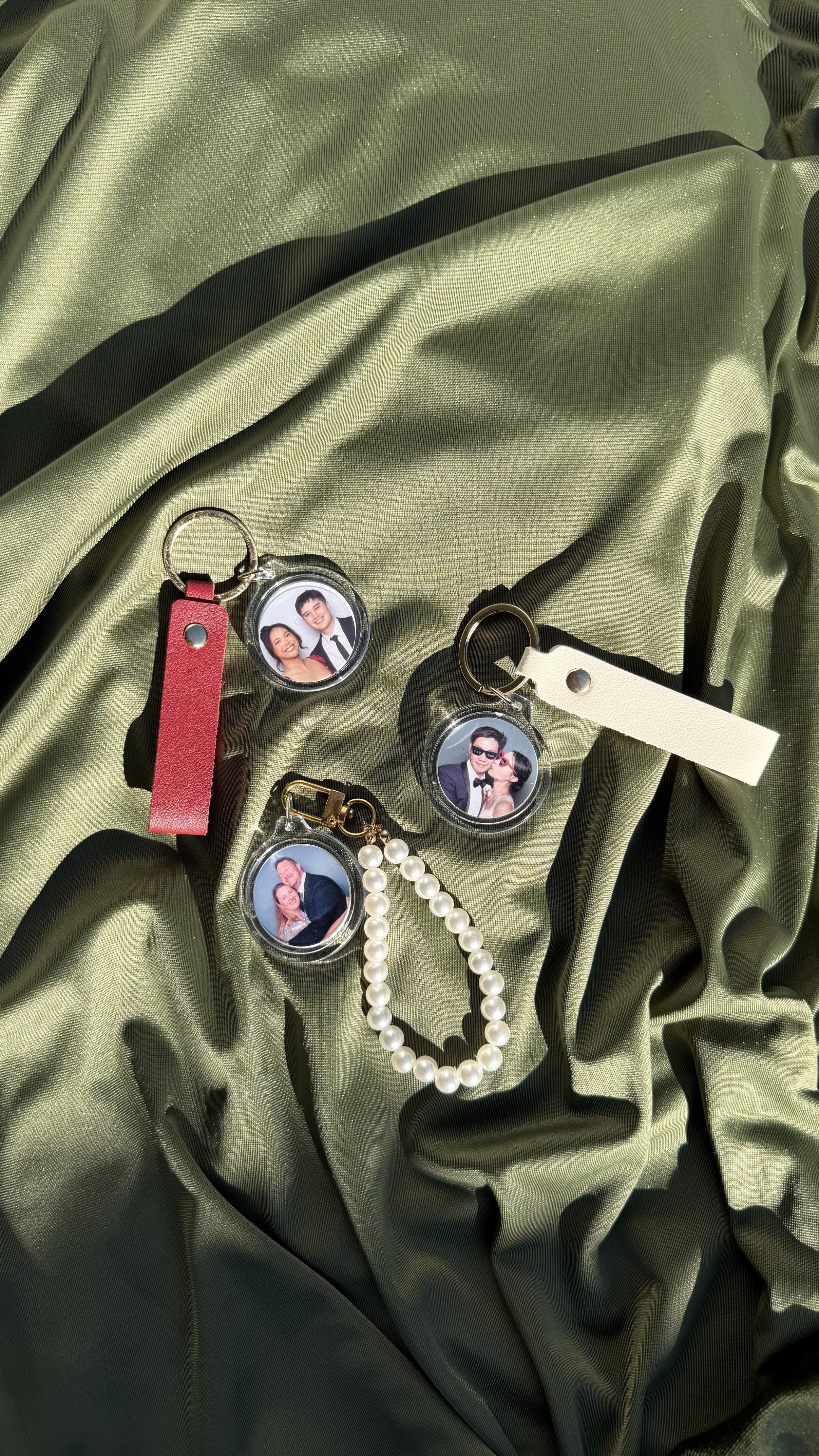 Fashion accessories with framed photos, including keychains, a pearl necklace, and leather tags, displayed on a shiny green fabric.
