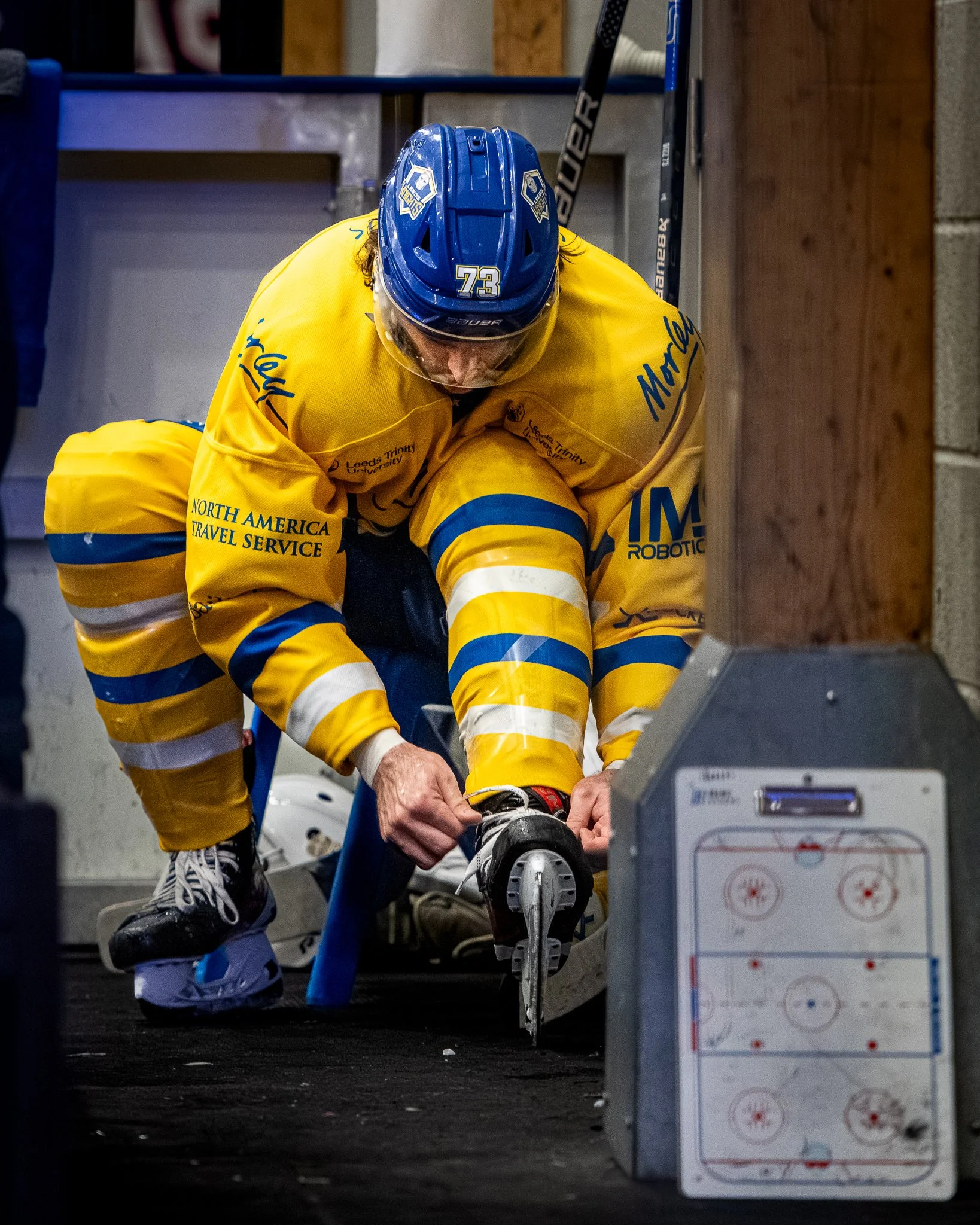 An ice hockey player wearing a blue helmet and a yellow jersey with merchandise logos is tying his skates in a locker room, with a hockey stick and hockey equipment nearby.