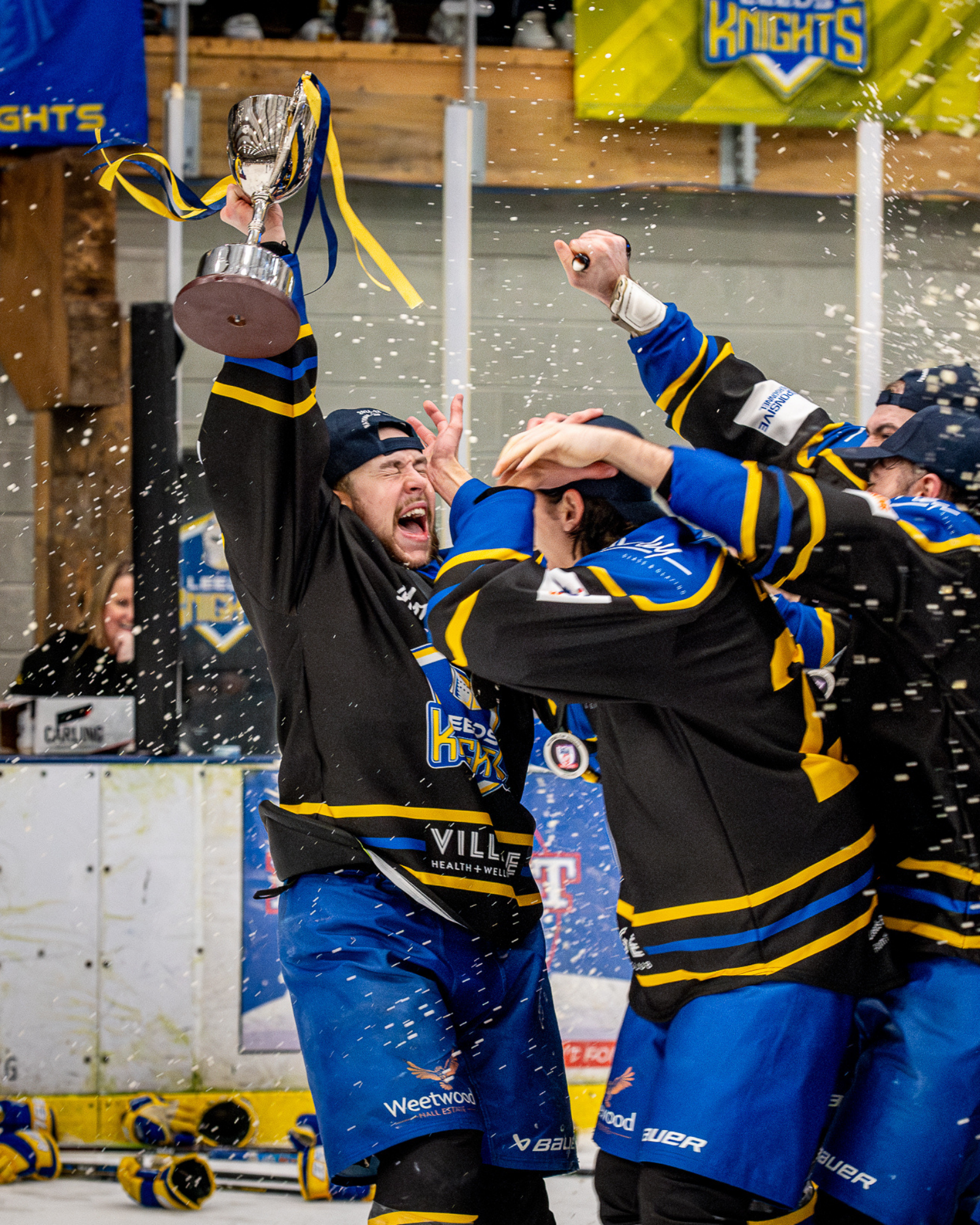 Hockey players celebrating on the ice after winning, with one holding a trophy and spraying champagne, in an indoor hockey arena.