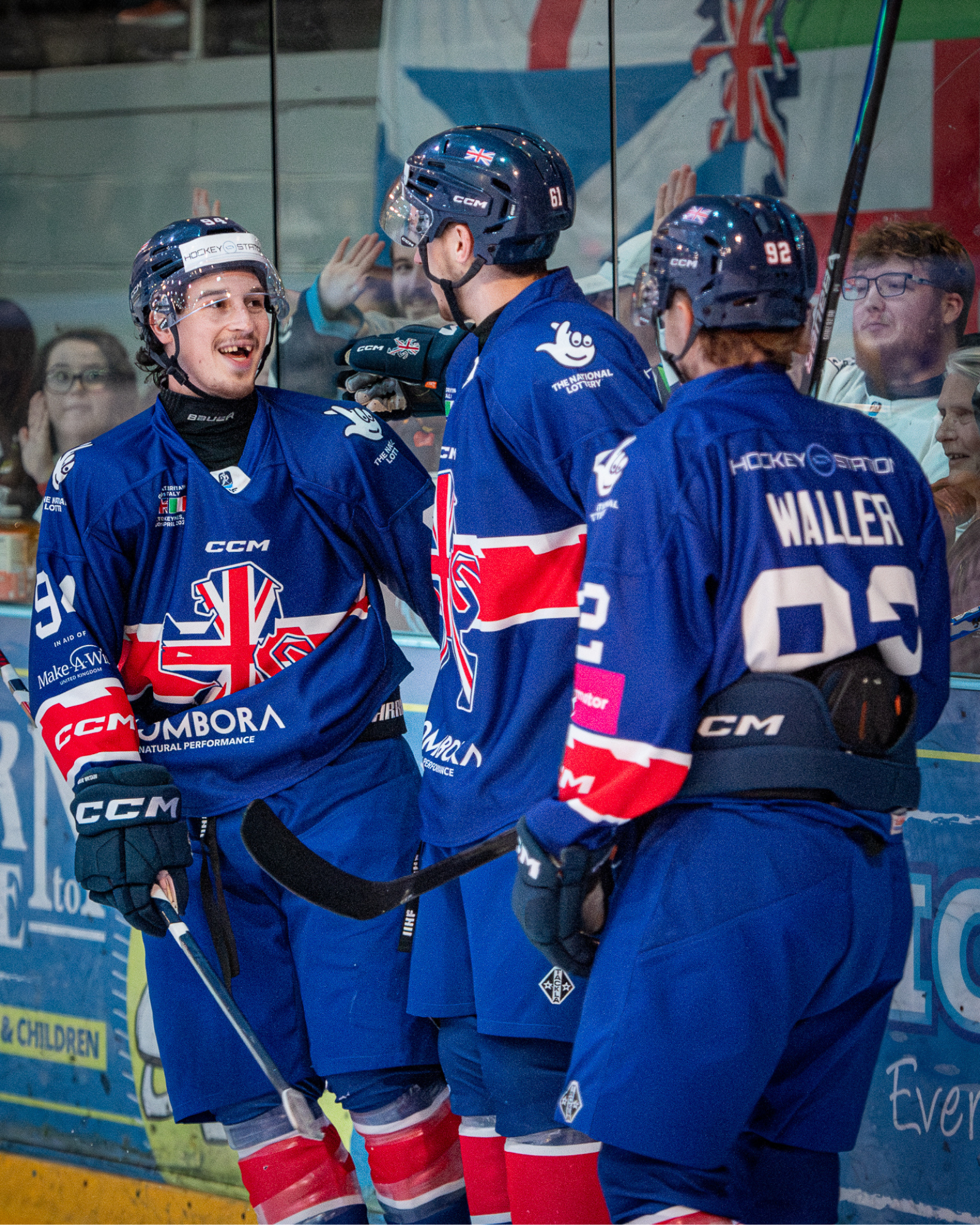 Ice hockey players in blue jerseys with a Union Jack design, celebrating during a game while standing beside the rink's glass.