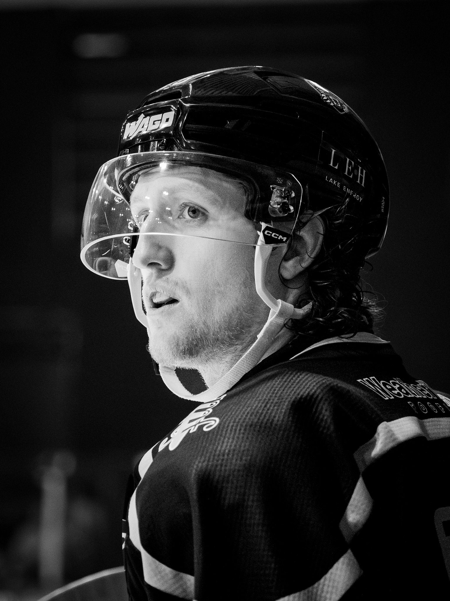 Close-up black and white image of a hockey player wearing a helmet with a transparent visor, with a serious expression, looking to the side.