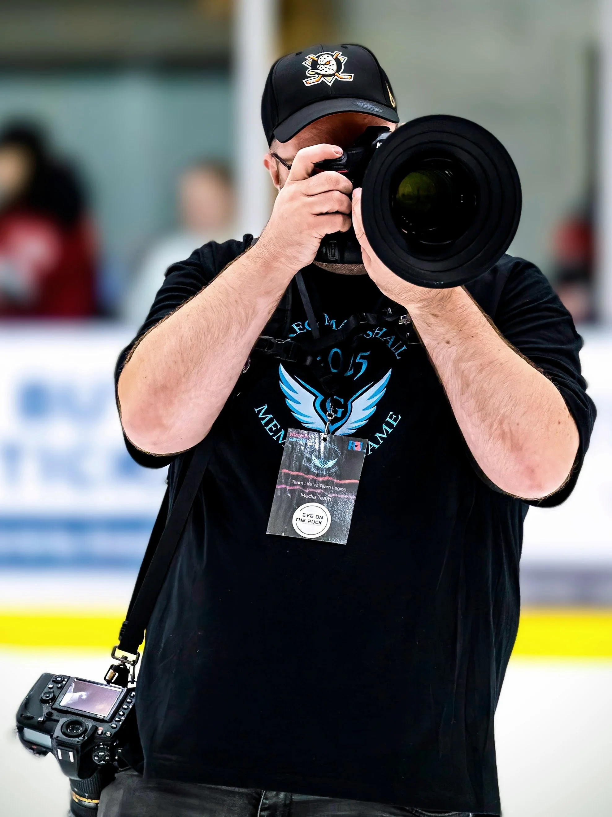 A person in a black shirt and baseball cap with a hockey team logo, holding a professional camera with a large lens, and wearing a media badge.