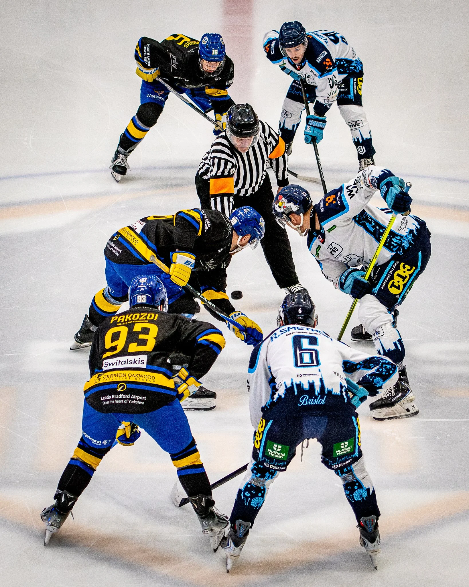 Two ice hockey players are face-to-face battling for the puck along with a referee watching closely on the ice. One player is in black and yellow, the other in white and blue.