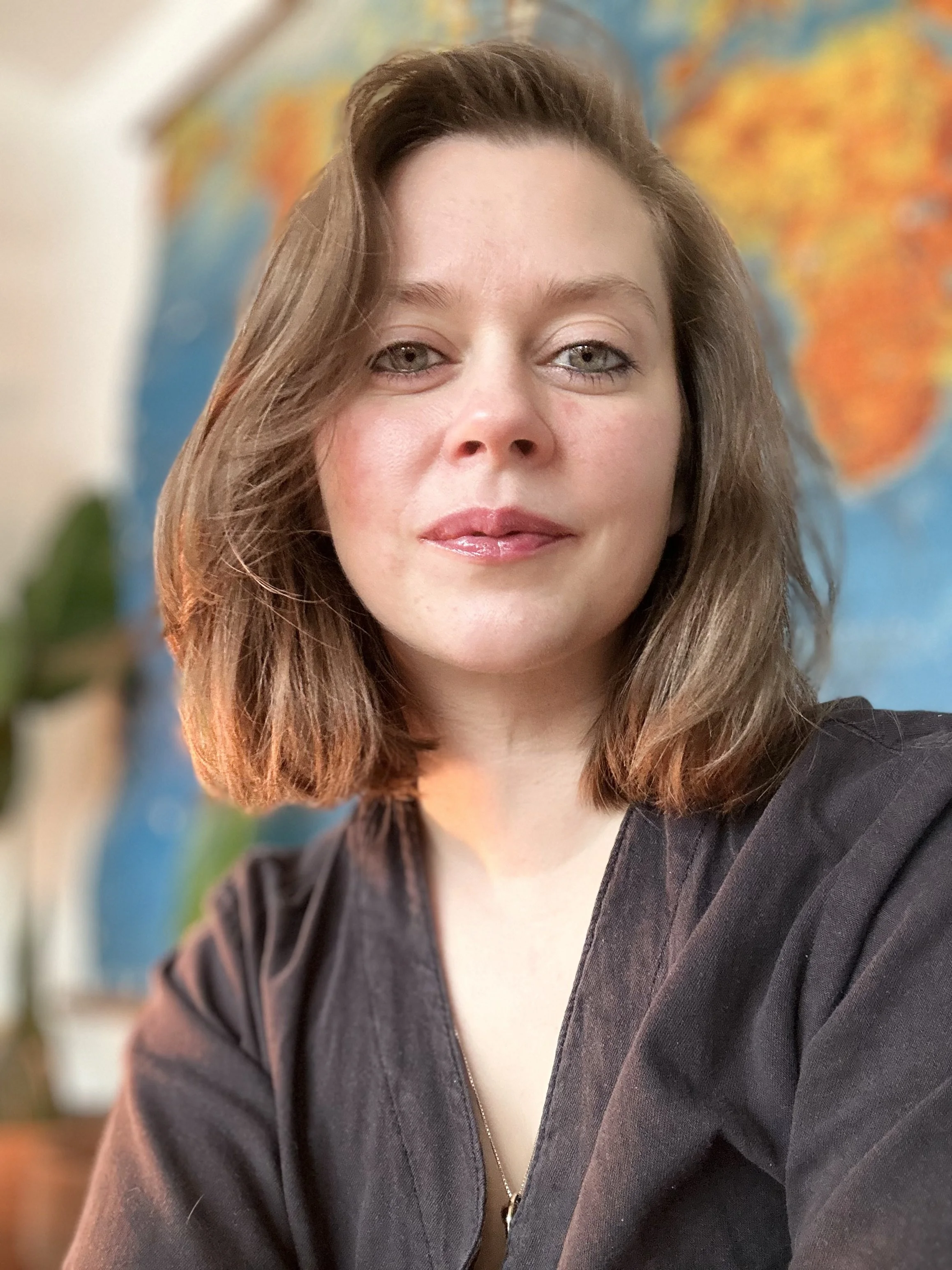 Nærbilde av en kvinne med brunt hår, smilende, i en svart jakke, med et fargerikt verdenskart i bakgrunnen.