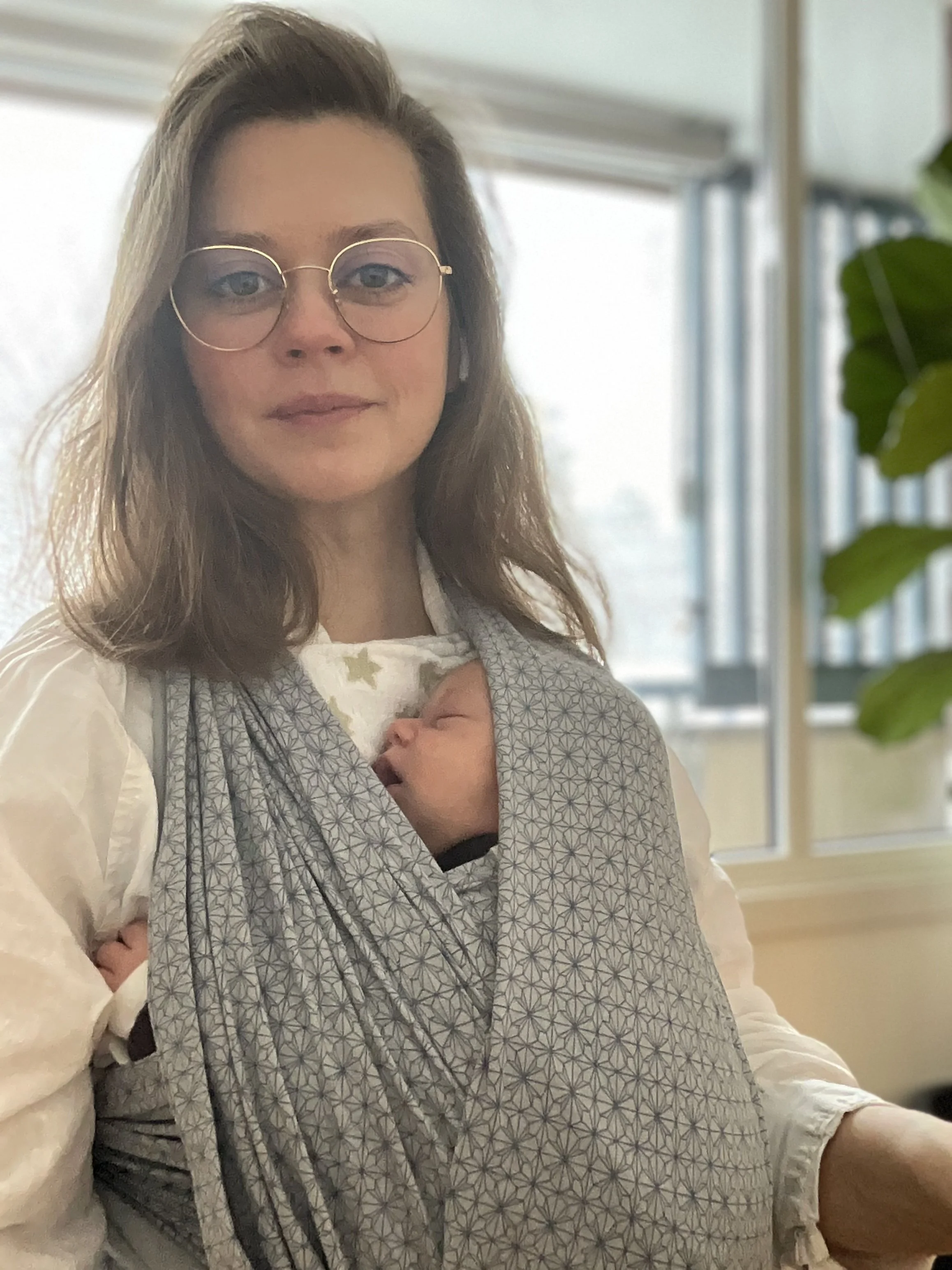 Kvinne med brunt hår, iført rund, gullfarget brille, som holder et barn i en bæreslynge. Det er et rom med store vinduer og innendørs planter.