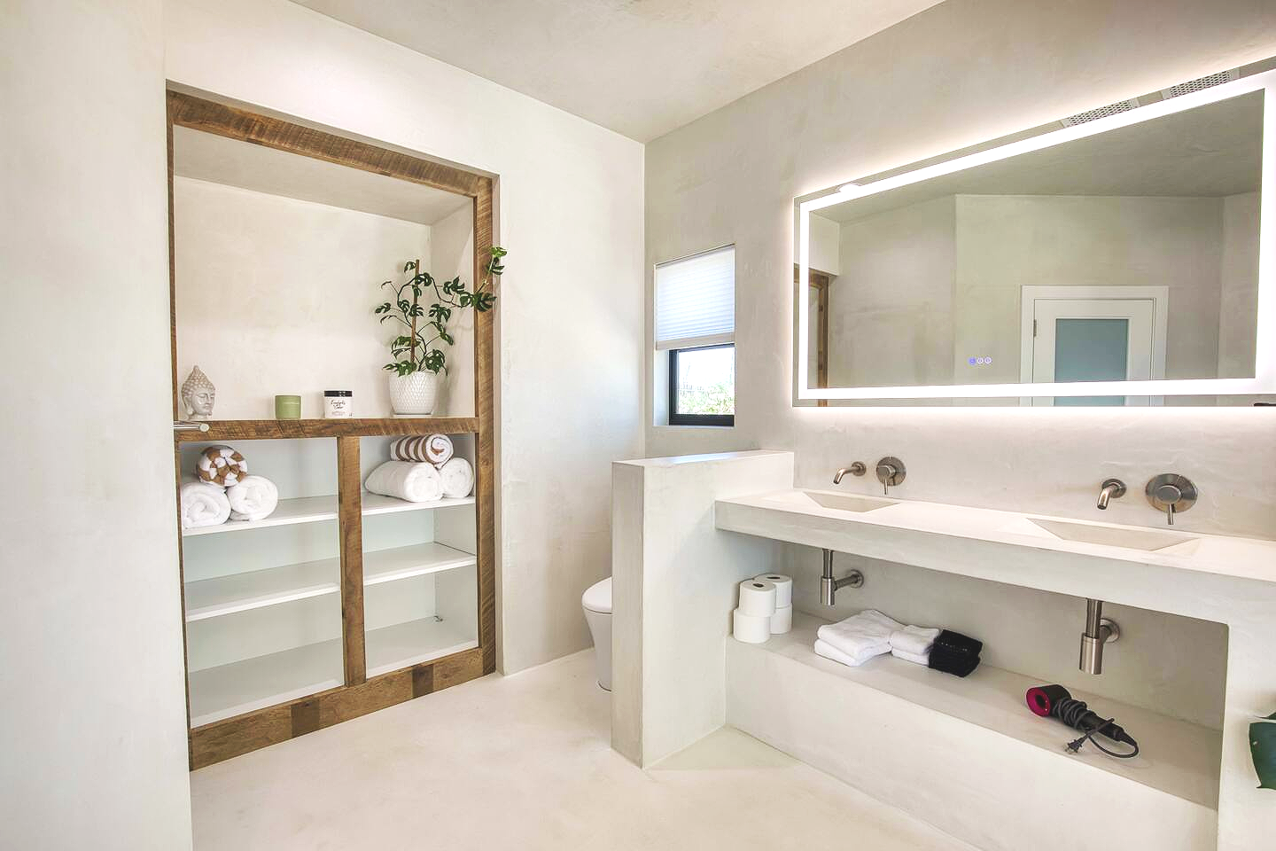 Modern bathroom with white walls and countertops, large illuminated mirror above dual sinks, small window, open shelving with towels and decorative items, and a toilet separated by a partial wall.