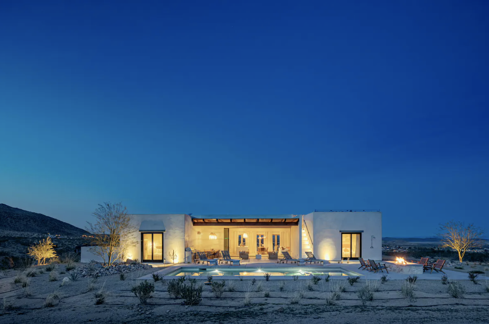 Modern house with a backyard pool and outdoor seating, illuminated at night, with desert landscape and mountains in the background.
