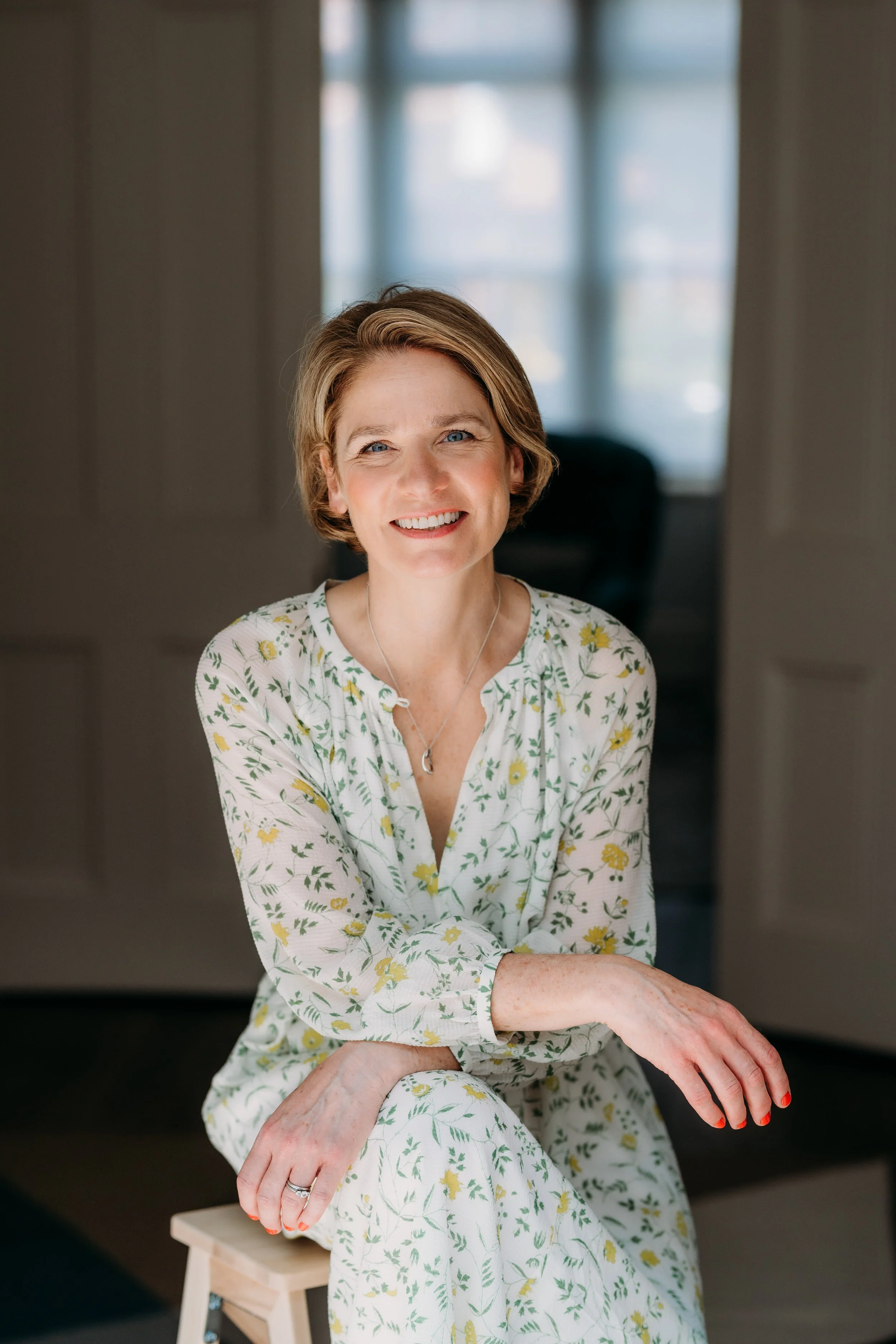 Victoria Bain, Founder of Victoria Bain Embroidered Textiles.A smiling woman with short light brown hair, wearing a white dress with yellow and green floral pattern, sitting in front of a window with sheer curtains, with her hands resting on her lap.