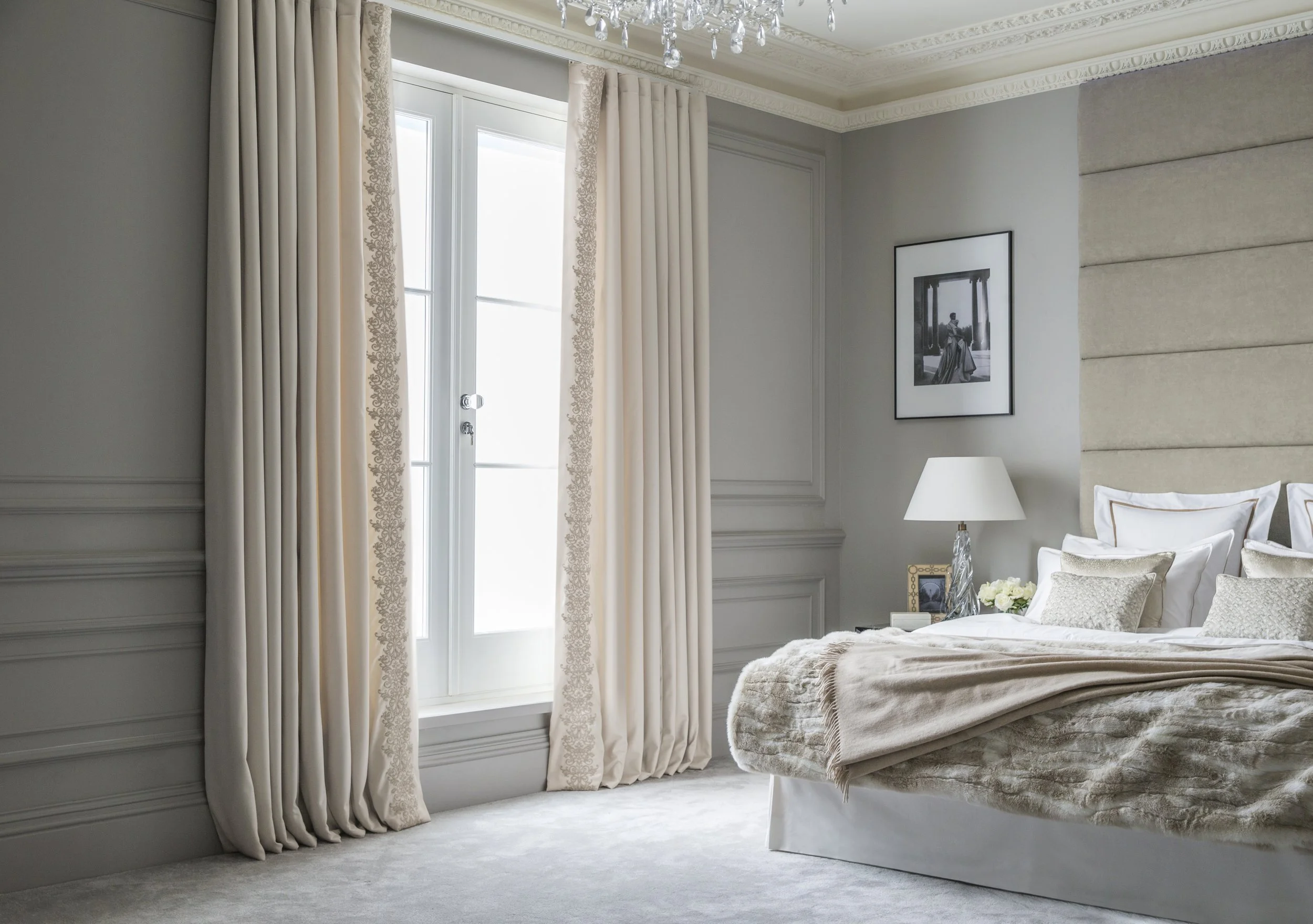 A bedroom with large windows, cream curtains, a framed black and white photo on the wall, a white lamp, and a bed with a beige bedspread and decorative pillows.